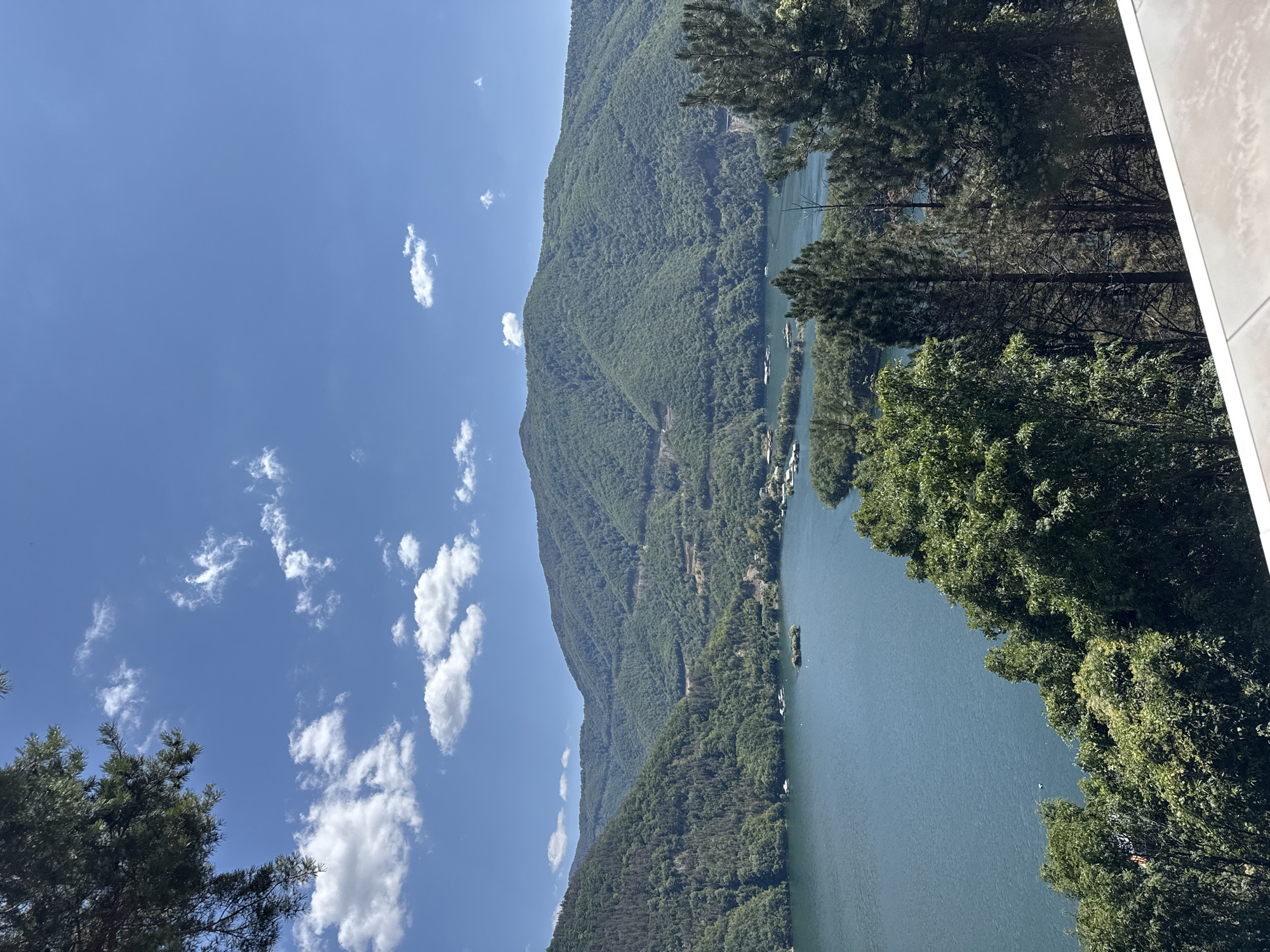 The image shows a scenic view of a lake surrounded by lush green mountains under a bright blue sky with scattered white clouds. In the foreground, there are trees, and in the background, the mountains are densely forested. The lake has a calm surface with a few small islands and some structures, possibly houses or boats, along its shores and on the water. The overall impression is one of a serene natural landscape. This view is consistent with the scenery found in and around Mihalkovo, Bulgaria, known for its natural beauty and mountainous terrain. There are no people visible in the image.