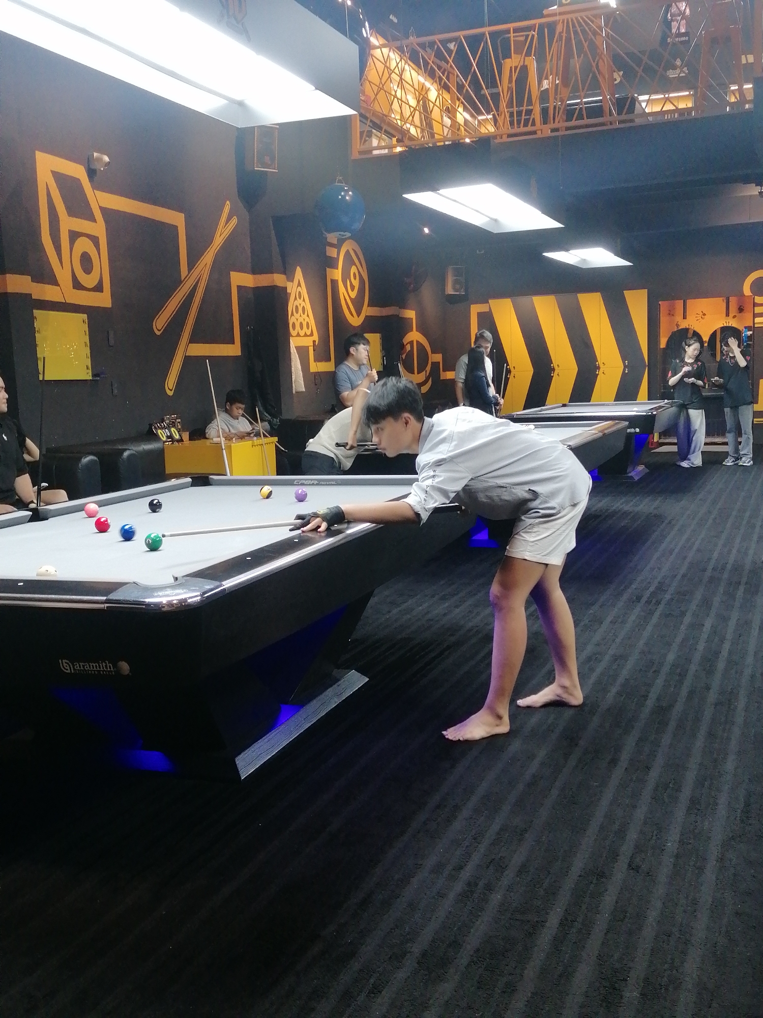 The image depicts an active indoor pool hall in Da Lat, Vietnam, characterized by a modern and energetic ambiance, likely during evening hours given the artificial lighting.
In the foreground, a young man, barefoot and dressed in a light grey short-sleeved shirt and white shorts, leans intently over a black-framed pool table to execute a shot. His left hand, wearing a black glove, forms a bridge for the cue stick. On the grey felt surface of the table are several colored pool balls (red, green, black, purple, yellow) and a white cue ball. The table's side displays the brand "Aramith Billiard Balls," and its base is illuminated with subtle blue LED lighting.
The background reveals several other individuals engaged in various activities. To the left, a man in a black shirt sits on a black sofa. Behind the foreground table, a young boy sits on another black sofa, appearing to look at a phone, near a yellow box scoreboard displaying "0:0." Further back, more people are seen around other pool tables. One person stands near a table marked "CYGR ROYAL," holding a cue. In the far right, two individuals, a man and a woman, stand beside a pool table, both focused on their phones.
The hall's interior design is distinctive, featuring dark grey or black walls adorned with bright yellow geometric and abstract patterns, including shapes resembling a cube, crossed cue sticks, a billiard triangle, and a '9' ball. A section of the wall in the mid-right features lockers with bold yellow and black diagonal stripes. Overhead, long rectangular fluorescent lights provide illumination, and a large yellow metal truss structure, possibly a mezzanine or decorative element, spans the upper background. A blue spherical object hangs from the ceiling, and a speaker is mounted on a wall. The floor is covered with dark grey striped carpeting. The overall scene conveys a casual and social atmosphere for playing billiards.
