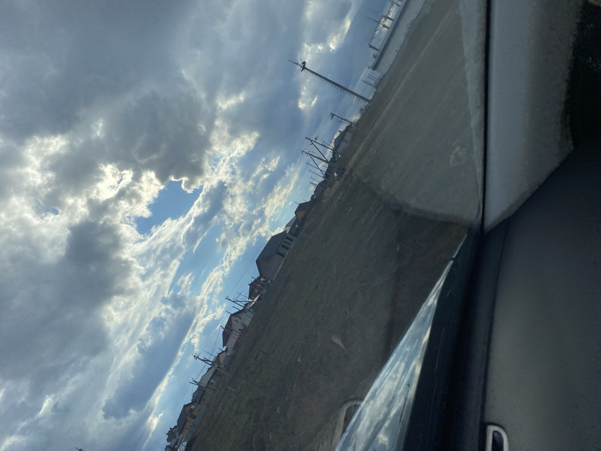 The image presents a dynamic outdoor scene, captured from the interior of a vehicle, likely in Chaikovsky, Russia. The sky dominates the upper portion of the frame, showcasing a dramatic blend of heavy, dark grey clouds interspersed with bright patches of blue sky. Sunlight breaks through these openings, creating areas of intense brightness on the clouds.
Below the expressive sky, the landscape appears as an undeveloped or semi-developed area, possibly a field of dry grass or dirt, stretching towards a distant horizon. A row of residential buildings, possibly houses, with dark roofs, lines the horizon on the left side of the image. Numerous utility poles, carrying power lines, extend across the landscape, connecting to or passing by these structures. Towards the mid-right, a tall, slender communication tower or antenna stands prominently against the sky; a small, dark shape, possibly a bird, can be seen perched near its apex. Further to the right, a large, possibly industrial or structural element, appears to reflect the ambient light.
The right side of the photograph features the dark interior of a car, including part of a door frame and a window with reflections, confirming the perspective is from inside a vehicle. The time of day is clearly daytime, with bright sunlight indicating either late morning or afternoon. No specific human activity is discernible, and no visible text is present.