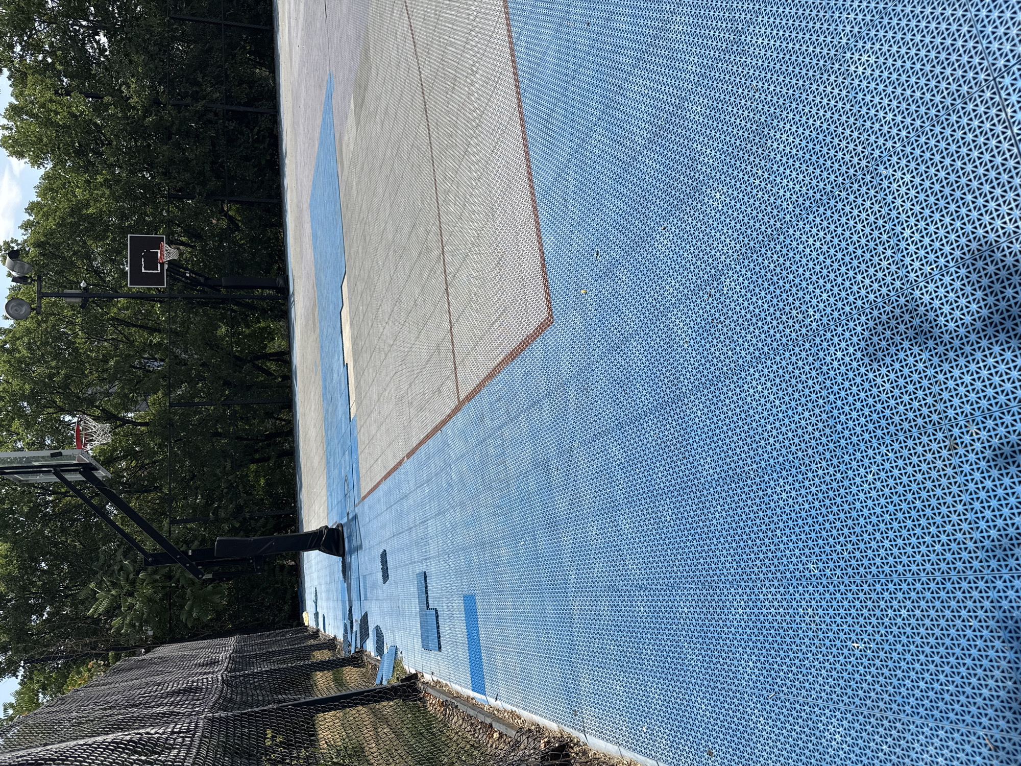 An outdoor basketball court with a blue interlocking tile surface is depicted. A chain-link fence runs along the left side of the court. A basketball hoop with a glass backboard is visible on the left, and another hoop with a dark backboard is in the background on the right. Trees and a bright sky form the background. The court appears to be partially under construction or renovation, with some blue tiles displaced near the fence. The scene is set in Yerevan, Armenia.