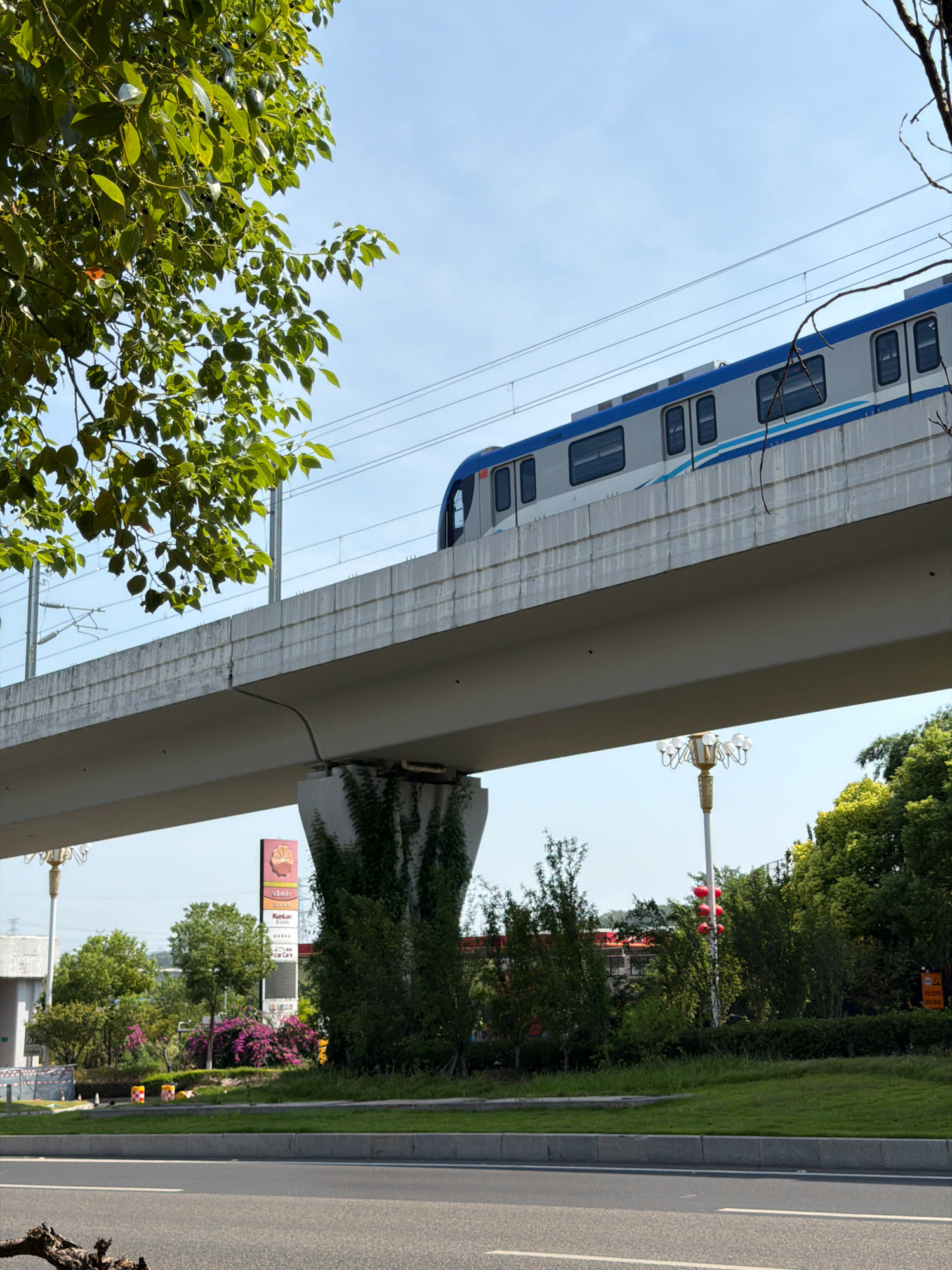 In Jiangjin, China, on a clear day, a light rail train travels along an elevated track. The train, with a white and blue exterior, is visible from the side, its windows reflecting the sky. The elevated track is supported by a concrete pillar covered in greenery. Below the track, a road runs parallel, bordered by a grassy median and trees. A tall sign with advertisements for "u smile" and "KirLan" is visible in the background, alongside a street lamp with decorative lights. The overall scene depicts urban infrastructure integrated with a natural environment.