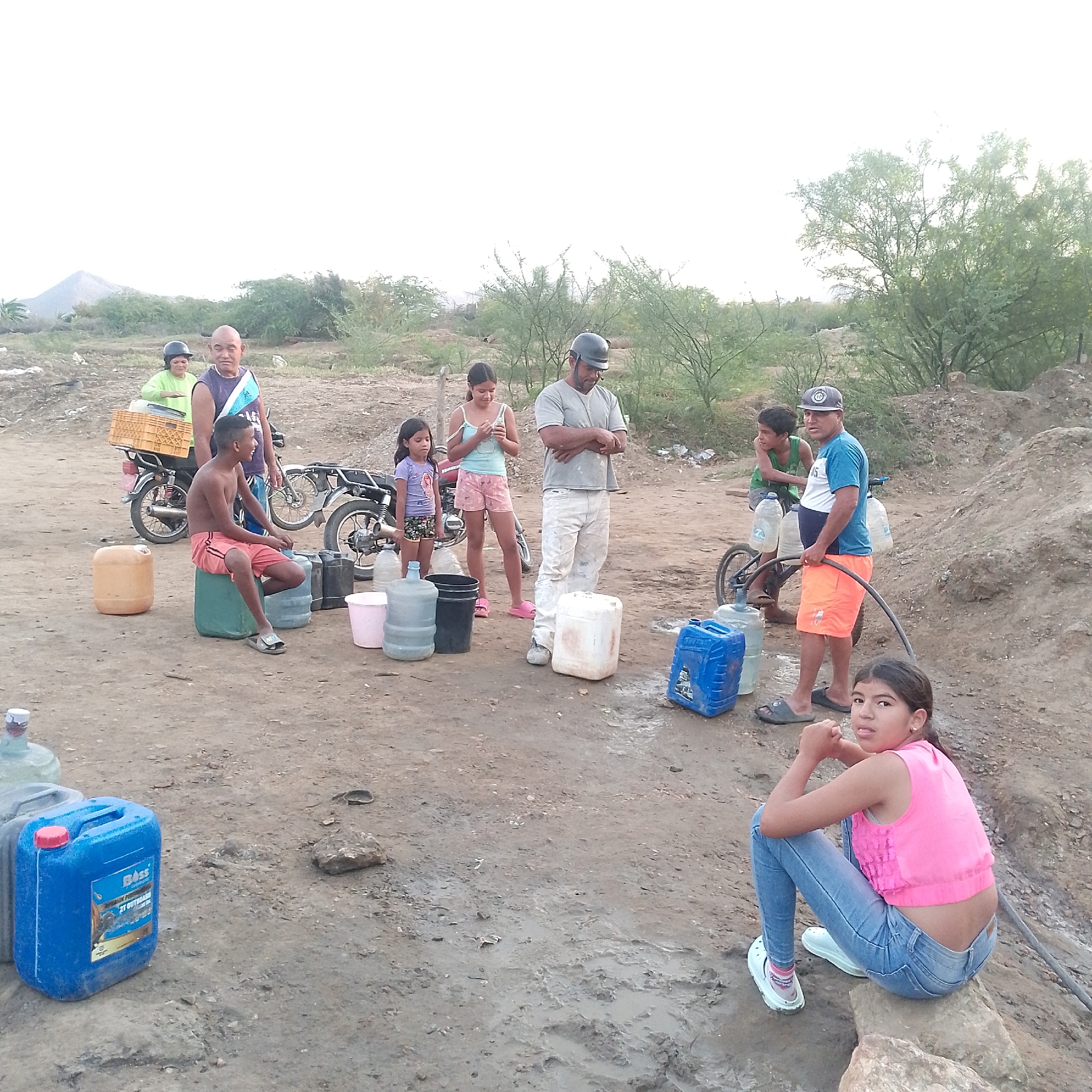 Several people are gathered in an outdoor area that appears to be in Altagracia, Venezuela.
There are at least ten people present, including children and adults. Two people are on motorcycles; one motorcycle has a crate attached to the back. Another person is on a bicycle, which is laden with water containers. Several water containers of various sizes and colors are scattered throughout the scene.
The ground is dry and dusty, with some muddy patches. The background features low-lying vegetation and small hills. The overall lighting indicates it is daytime.