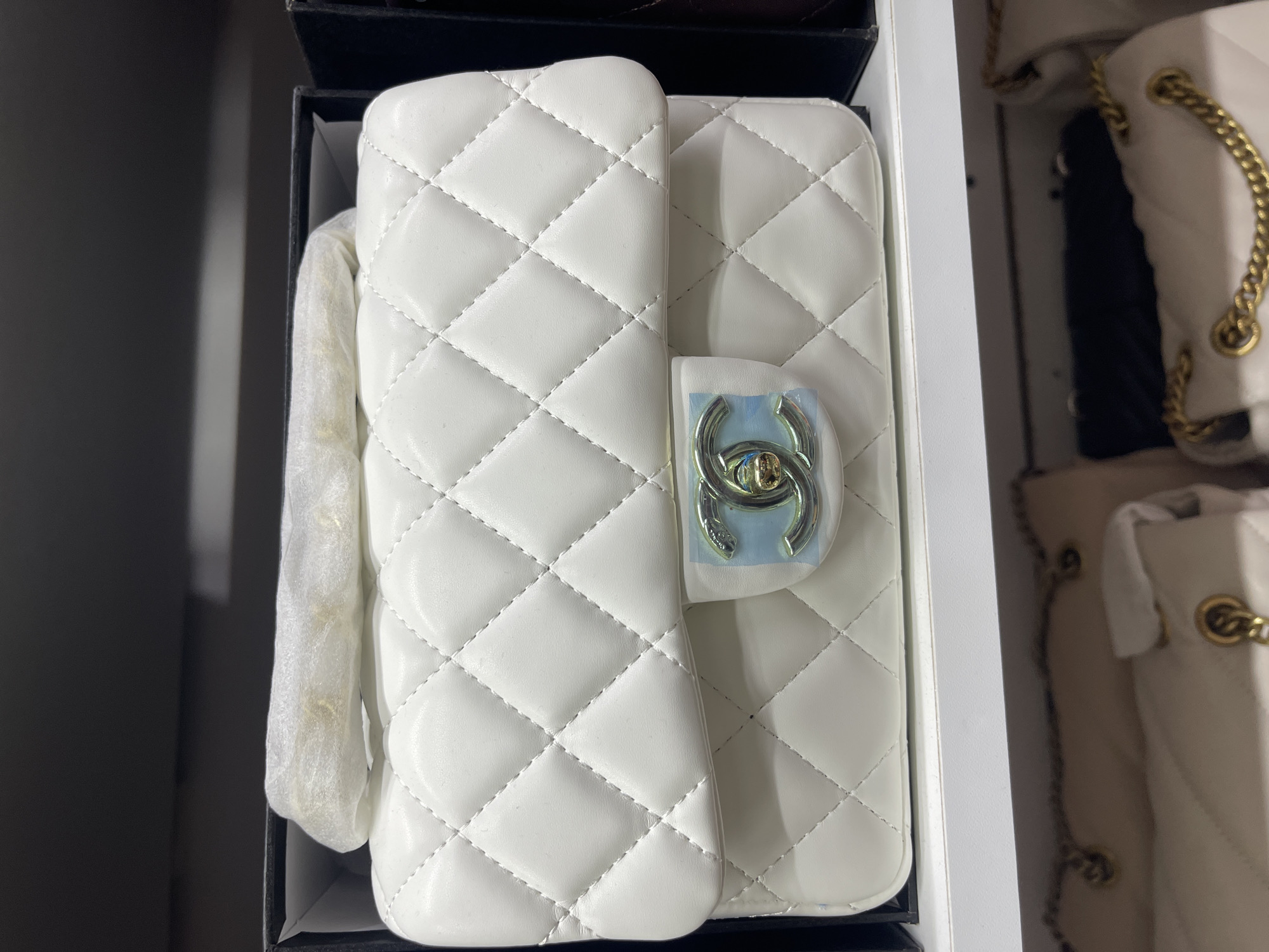 In a retail environment, possibly a store or a warehouse, a white quilted handbag with a metallic interlocking C logo clasp is displayed. The handbag appears to be new, as it is partially wrapped in translucent protective material and placed inside a black box. Below this handbag, on a white shelf, other handbags are visible, including one with a gold chain strap. The background suggests a display or storage area with multiple shelves and boxes, typical of a commercial setting. There are no individuals present in the immediate frame of the image. The scene is indoors, with artificial lighting.