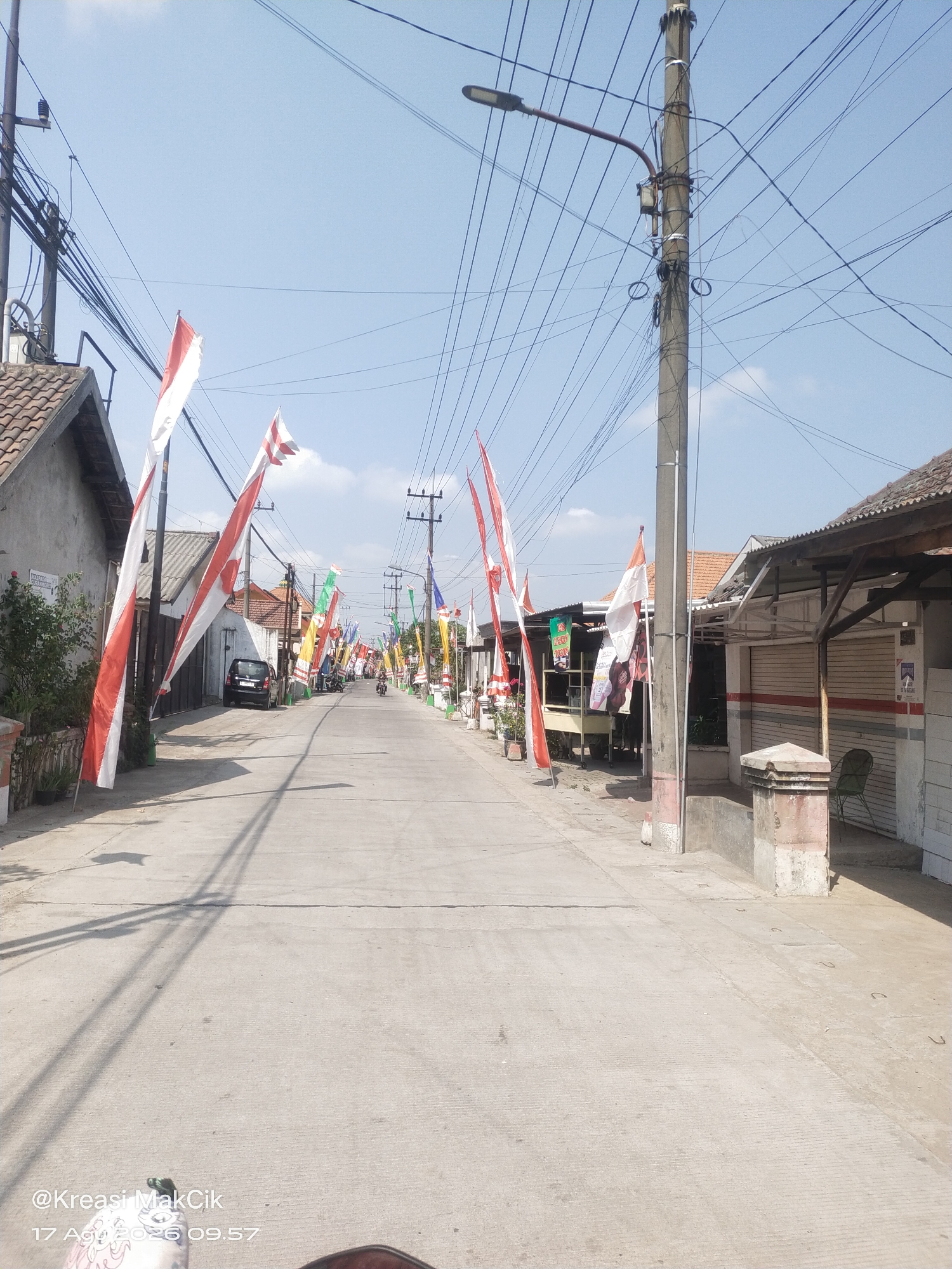 The image depicts a street scene in Sambi Bulu, Indonesia, decorated with numerous Indonesian flags. The flags, predominantly red and white, are hung along both sides of the asphalt road, indicating a festive or patriotic occasion, likely Independence Day. The street is lined with buildings, some appearing to be houses and others small shops or stalls, with their shutters down. A black car is parked on the left side of the road, and a motorcycle is visible further down the street. Utility poles with a dense network of electrical wires run along the street, casting shadows on the road surface. The sky is clear and blue, suggesting a sunny day. The overall environment is residential and commercial, with a clear emphasis on the celebratory decorations.