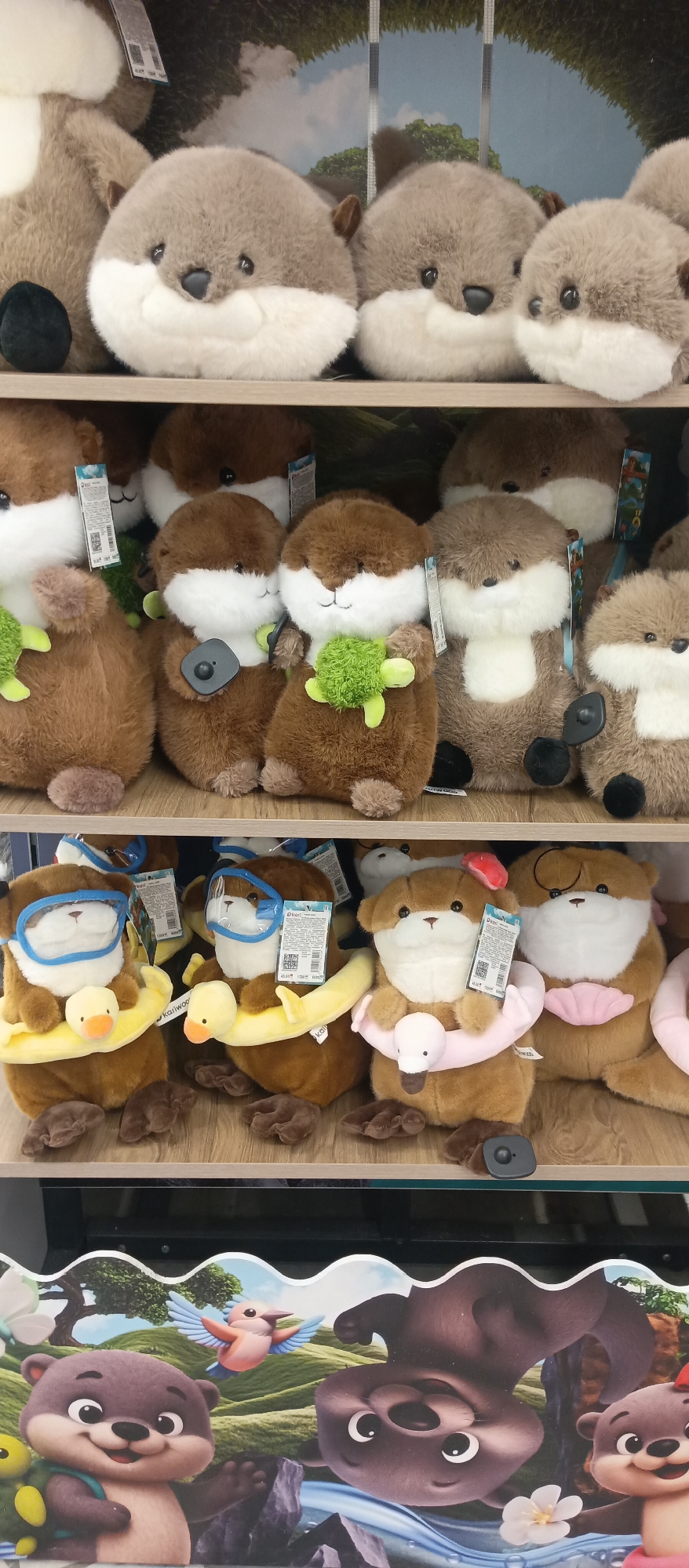 The image shows a display of otter plush toys on shelves in a store. The bottom of the frame shows a sign with animated otters. The location is Unknown, Unknown. The otters are displayed on three shelves. The top shelf features round, grey otters. The middle shelf features brown otters, some holding green turtles. The bottom shelf features brown otters wearing swimming accessories like goggles, floatation devices. The toys appear to be for sale.