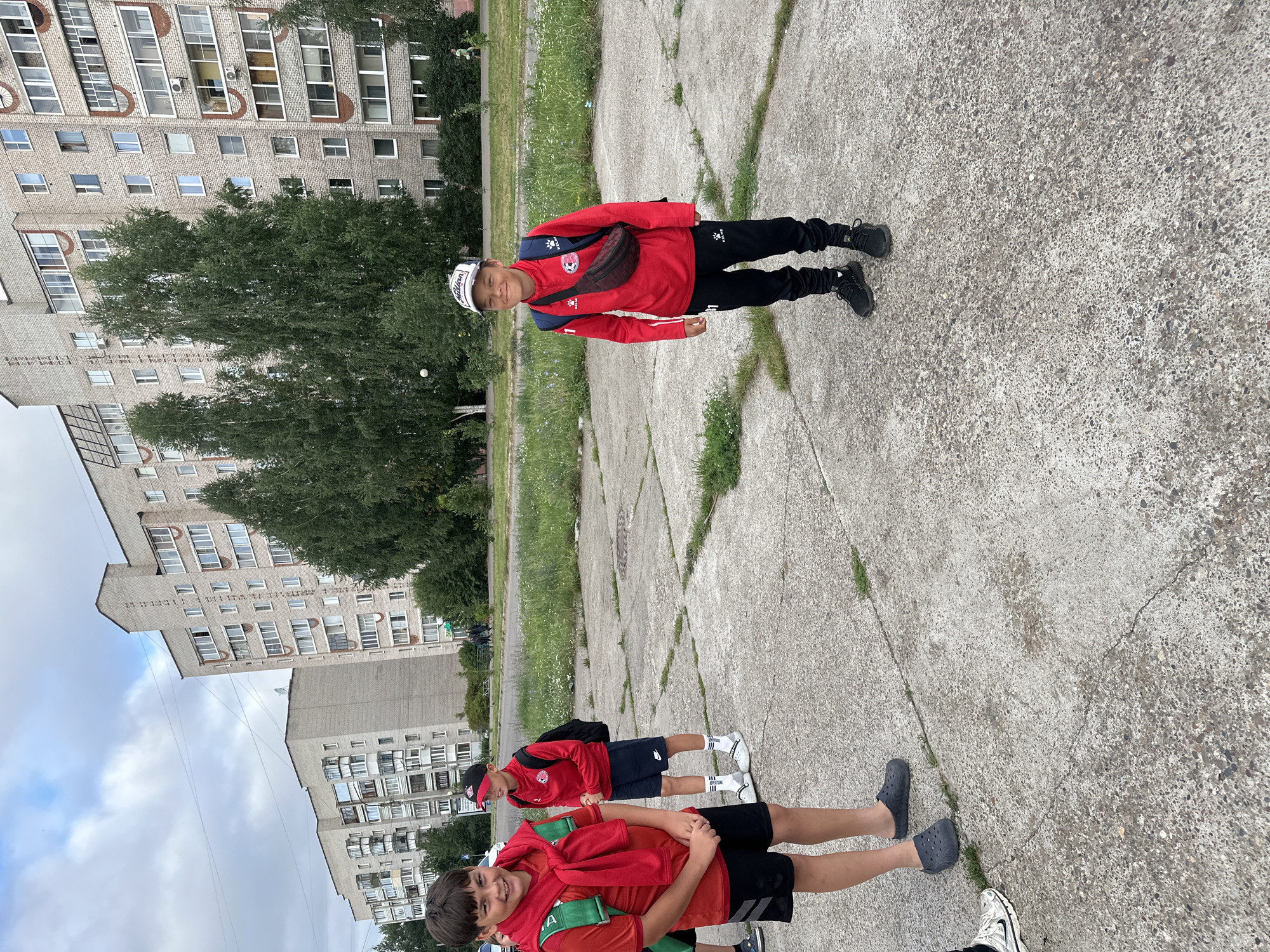The image depicts an outdoor scene in Chaikovsky, Russia. In the foreground, two young boys are standing on a concrete paved area. The boy on the left is wearing a red t-shirt, black shorts, and dark gray crocs. He has a red scarf draped over his shoulders and a green backpack. The boy on the right is wearing a red long-sleeved shirt, black pants, a black baseball cap, and black sneakers. He has a dark-colored fanny pack around his waist and a blue backpack. Behind the boys, a grassy area with trees is visible, and further in the background are multi-story apartment buildings. The sky is overcast.