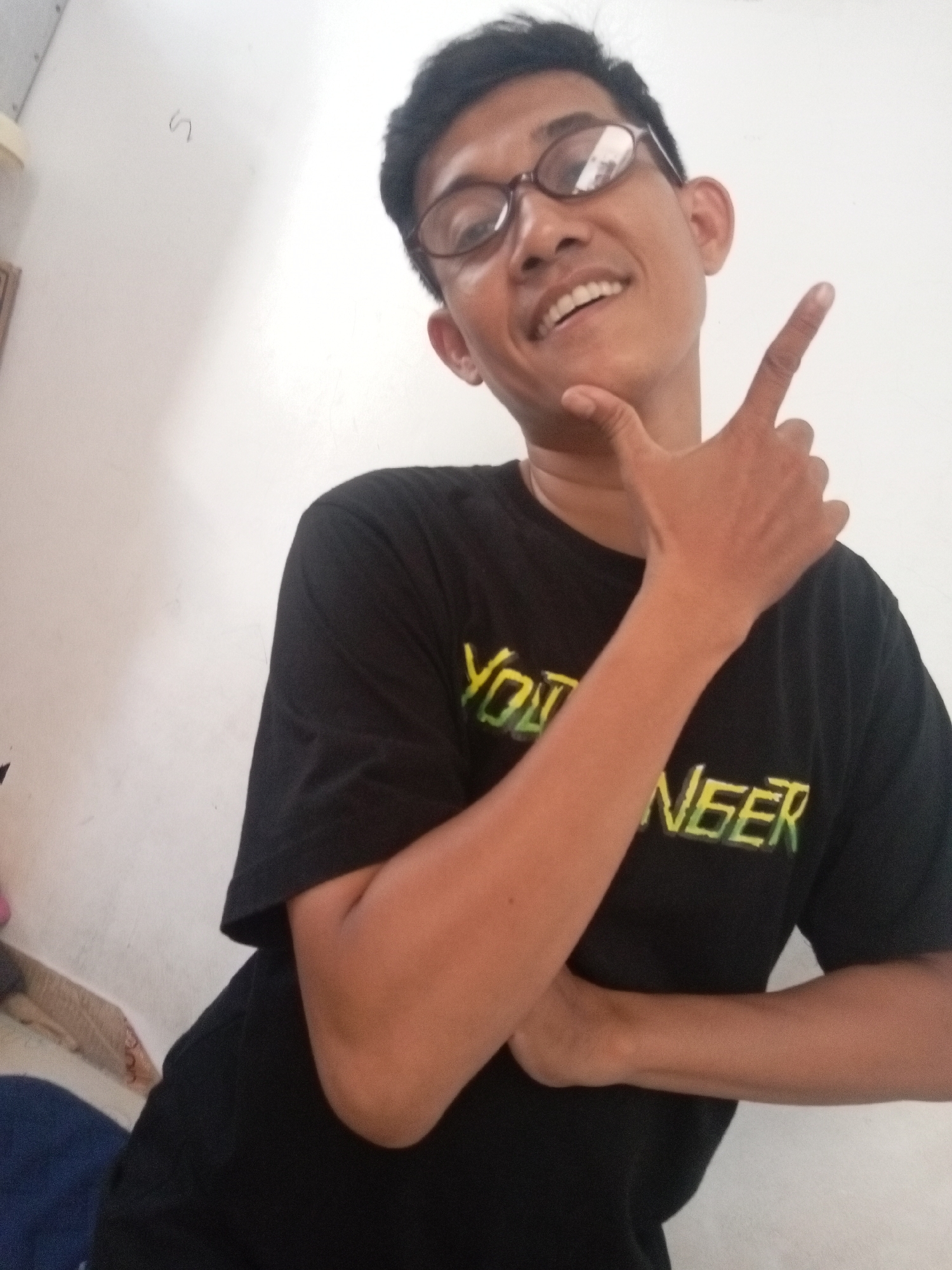A young adult male with dark hair and glasses smiles at the camera, pointing one finger upwards. He is wearing a black t-shirt with yellow and green text that appears to say "YOUNGSTER." He is standing indoors against a white wall. The lighting is neutral, and there are no discernible shadows. The image is a selfie, taken from a slightly low angle. The environment suggests a casual indoor setting, possibly a home or dormitory room. The image does not provide enough visual information to determine the specific location within Batam, Indonesia.
