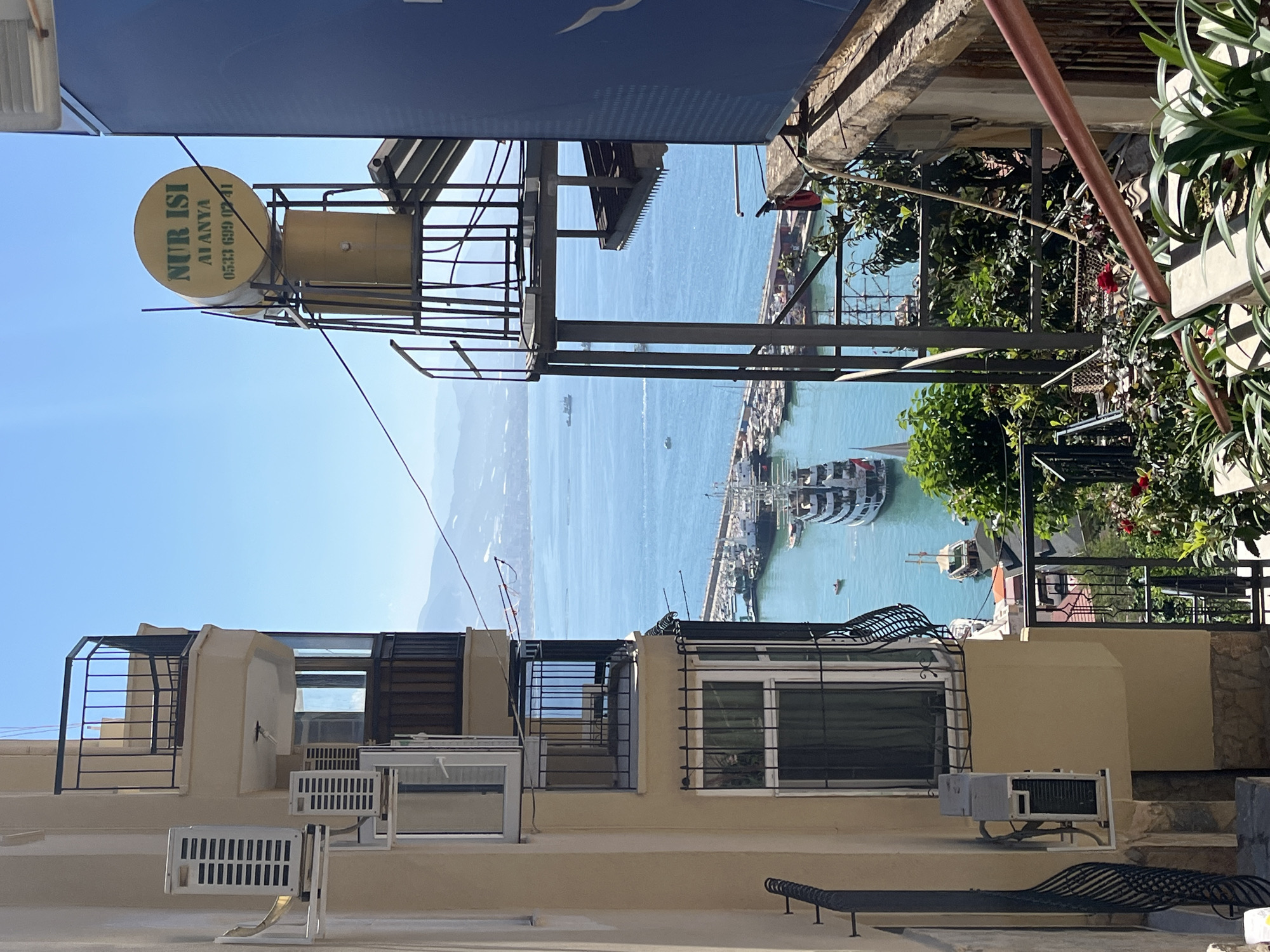 The image depicts a coastal scene in Alanya, Turkey, showcasing a partial view of the harbor with several boats and ships. The foreground is dominated by building rooftops and exterior structures, featuring air conditioning units, windows with security bars, and metal railings. A cylindrical water tank labeled "NUR ISI ALANYA 0533 699 0241" is mounted on a metal frame above. In the background, the turquoise-colored sea extends towards a distant coastline, with a clear blue sky overhead. A mix of residential and commercial structures is visible along the harbor, partially obscured by green trees.