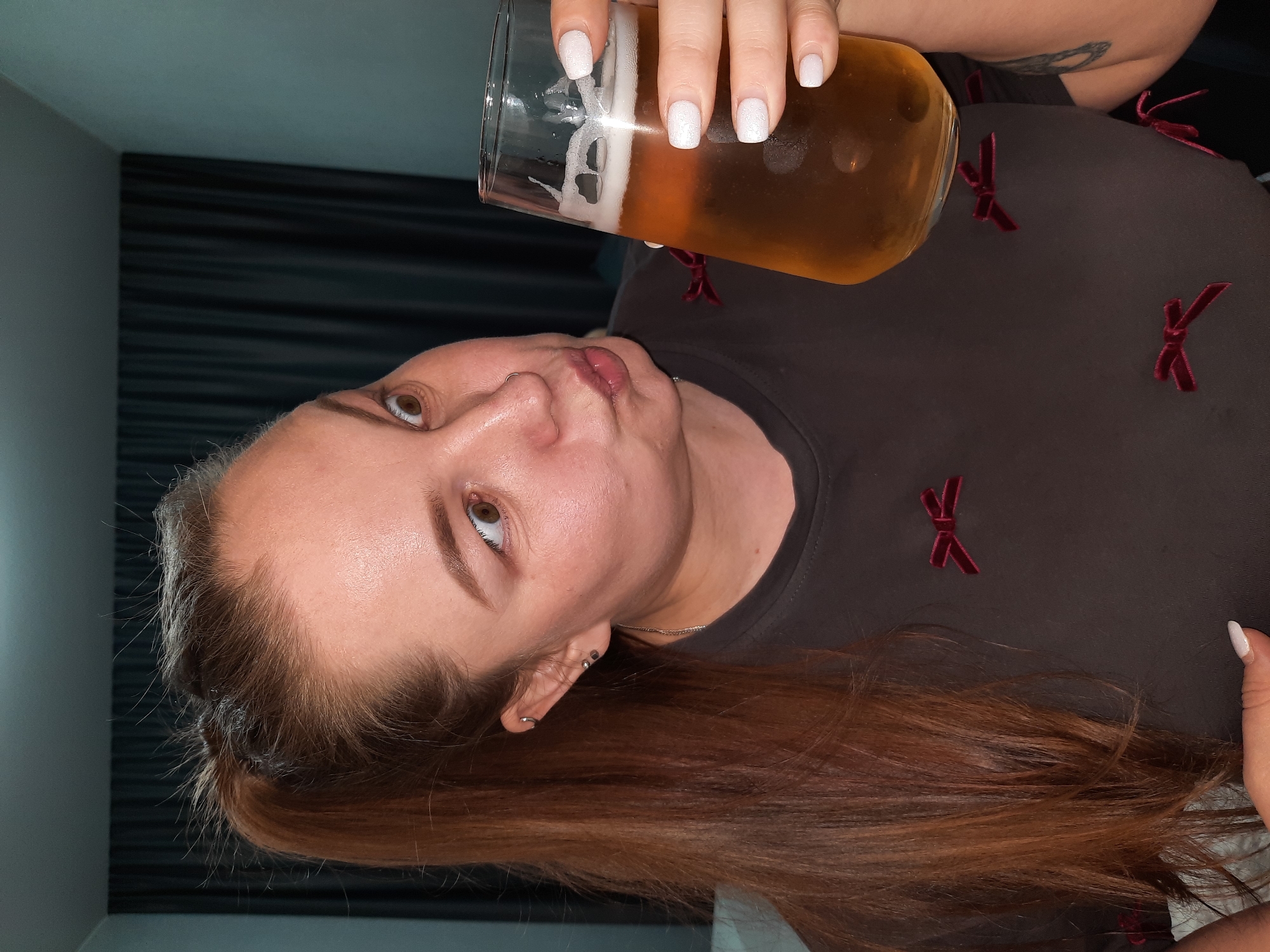 A woman with long, reddish-brown hair holds a glass of beer. She has light skin, brown eyes, and white manicured nails. She is wearing a dark gray shirt with small red bow accents. The background is out of focus, but appears to be a room with dark curtains. There is no visual information to suggest this image was taken in Pervomaysky, Russia.