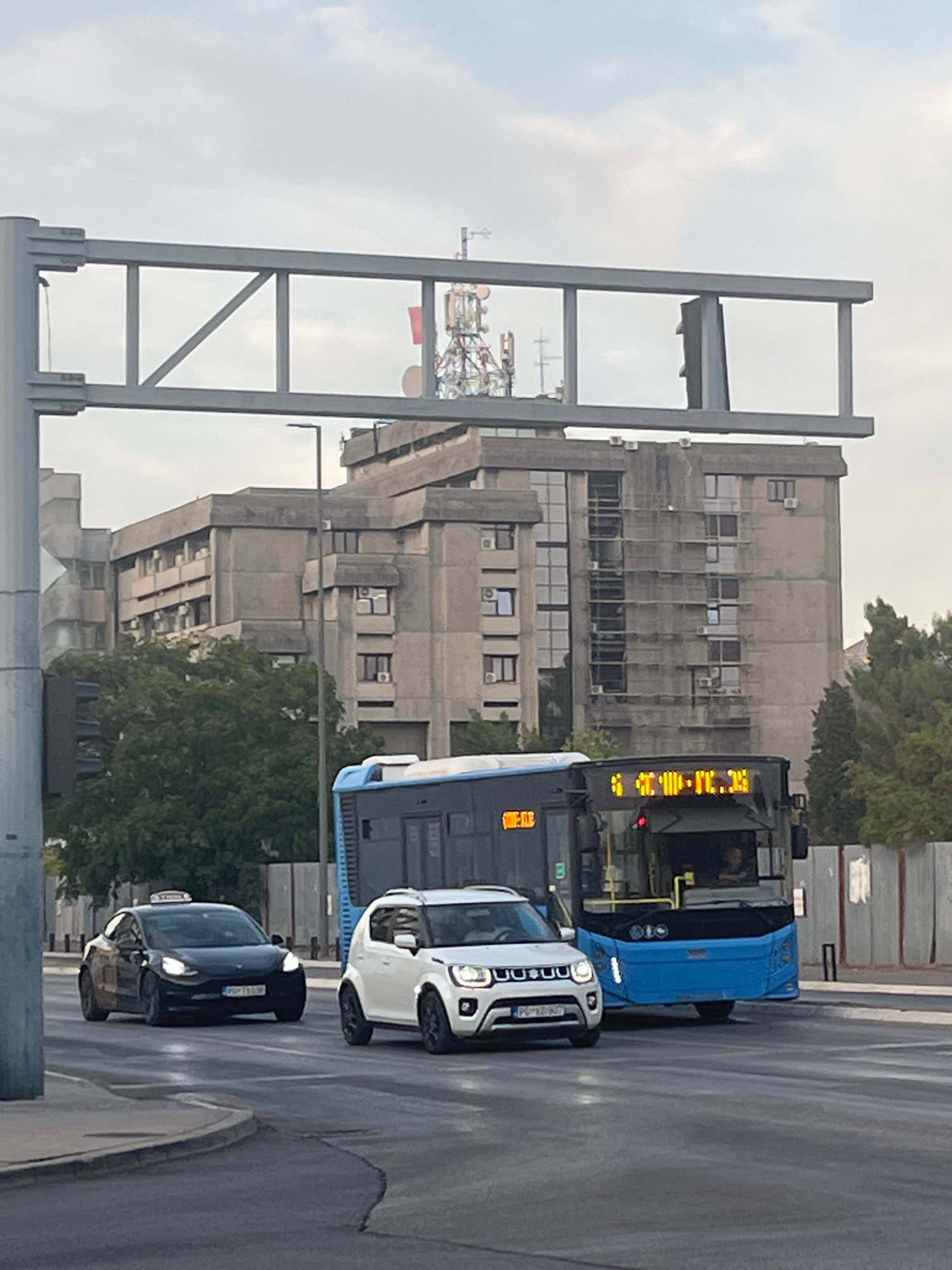 In Podgorica, Montenegro, on a cloudy day, a black Tesla Model 3 taxi with a visible license plate is driving on a wet road. A white SUV is beside it. Behind them, a blue bus is also on the road, displaying a route number in yellow digital text. In the background, a large, Brutalist-style concrete building with numerous windows and air conditioning units stands under a sky with multiple telecommunication towers. A traffic light structure is prominent in the foreground, with a gray metal frame overhead.