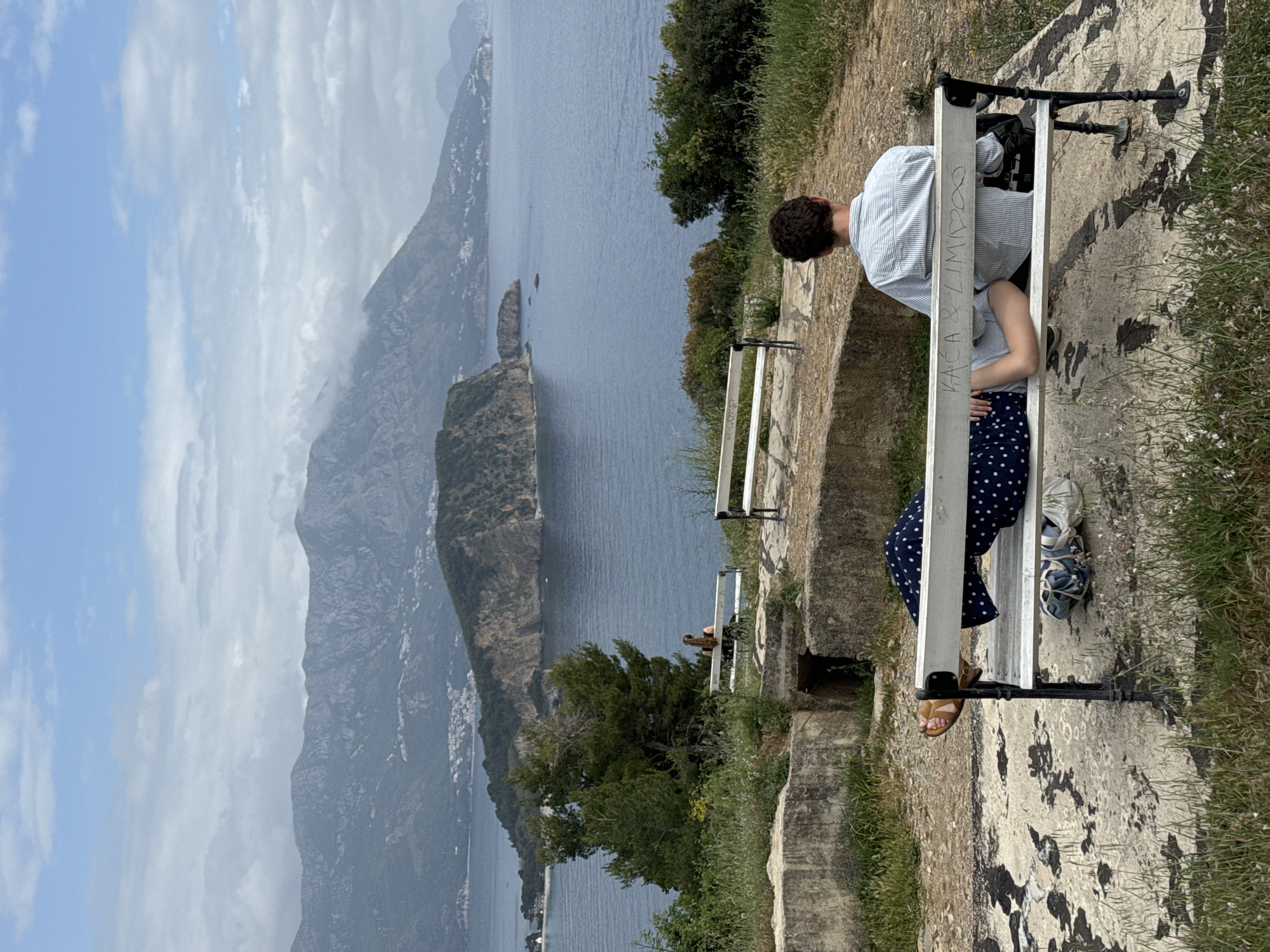 A man and woman sit on a white bench facing the Adriatic Sea and mountains near Budva, Montenegro. The man has short dark hair and wears a striped shirt. The woman is in a gray shirt and blue polka-dot skirt. They are on a stone path with grass growing on the edges. Another white bench is nearby. In the distance are mountains partially obscured by clouds.