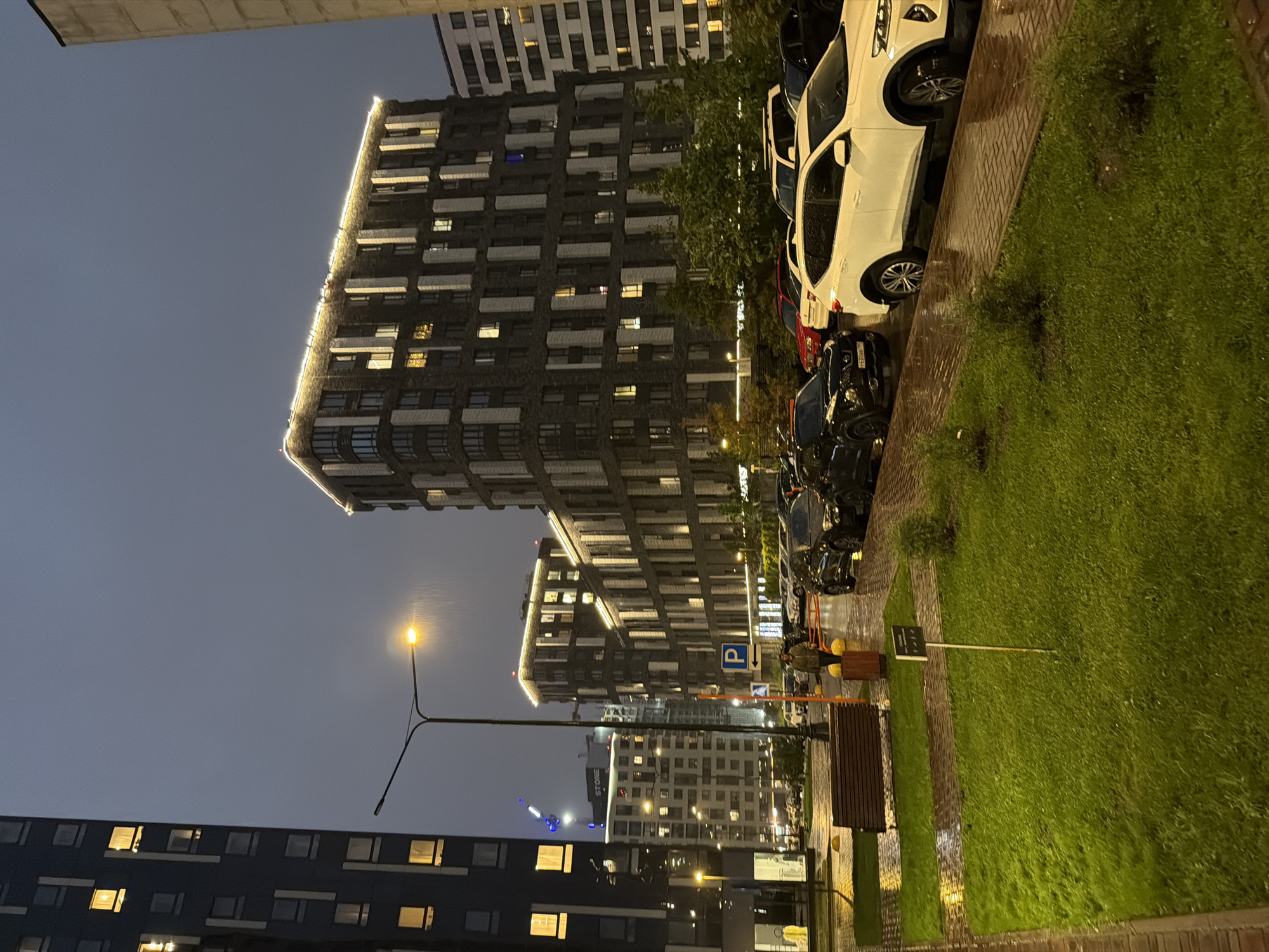 This image depicts a residential complex in Moscow, Russia, during nighttime and rainfall. The scene is illuminated by streetlights, building lights, and decorative lights along the top of one building, casting a yellowish glow on the wet pavement and grass. Tall, modern apartment buildings dominate the background, with multiple windows visible, some lit from within. In the foreground, a wet grassy area with small bushes is situated next to a brick pathway. A wooden bench is placed on the path. Several cars are parked along the street, their surfaces reflecting the ambient light due to the rain. A man in a green jacket is standing on the wet pavement near a parking sign. The sky is dark and overcast, consistent with rainy weather.