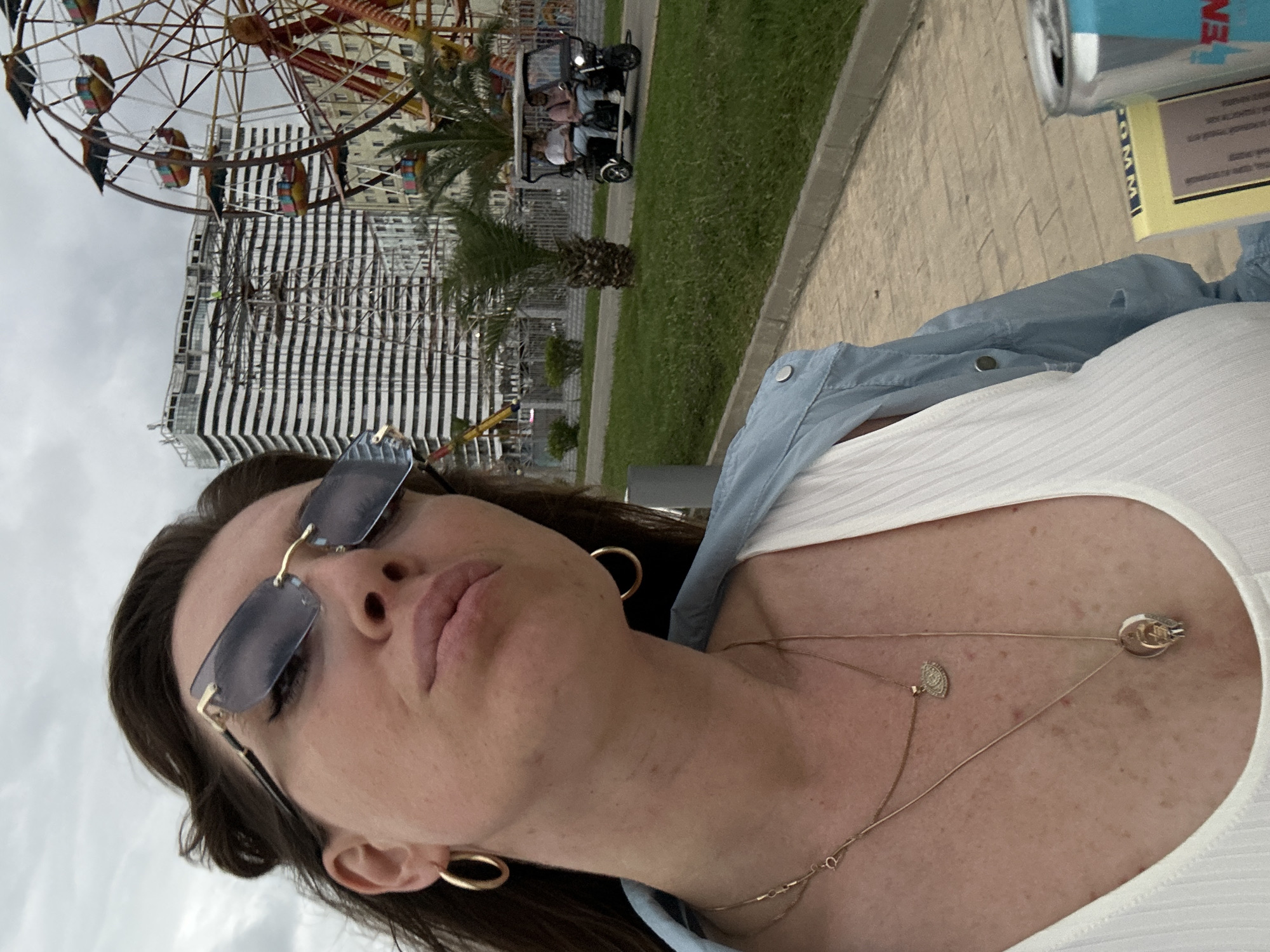 The image shows a woman taking a selfie with a Ferris wheel and a modern building in the background, likely in the city of Batumi, Georgia. She is wearing sunglasses, hoop earrings, and layered necklaces. In the background, a small electric vehicle with two passengers drives along a paved path. The area appears to be a promenade or park, with grass and palm trees. The building in the background is under construction, with scaffolding visible. A can and a book are placed on a surface in the foreground.