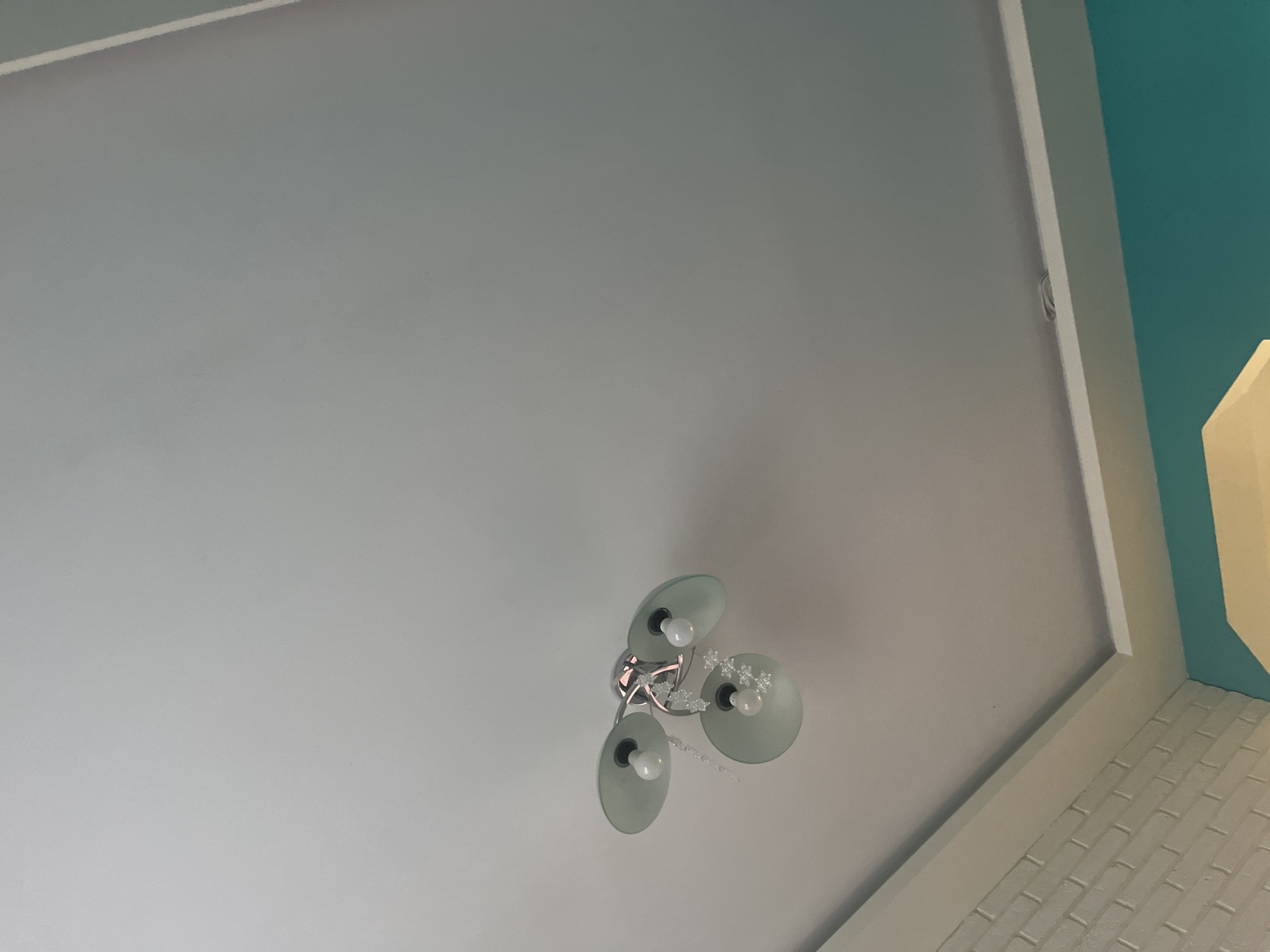 The image depicts a ceiling-mounted light fixture with three frosted-glass shades, each containing a light bulb. The fixture is attached to a white ceiling. The corner of the room is visible, showing white trim and a wall painted in teal and yellow. A section of brick wall is seen along the ceiling. The location of the image is Pokrovnik, Bulgaria.