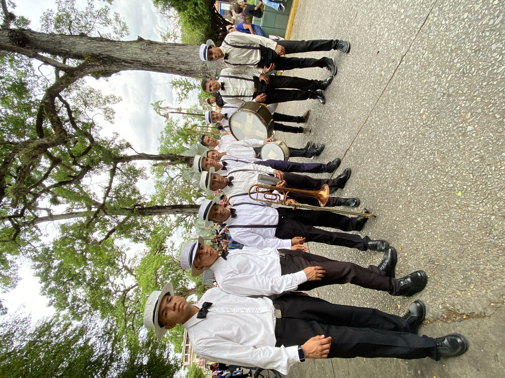 A band in Tinaquillo, Venezuela, stands in a line on an asphalt street, beneath the canopy of a large tree. The band members wear matching outfits consisting of white shirts, black pants, black shoes, and white hats with black bands. They are also wearing bowties. Some band members carry musical instruments including a trombone and drums. A small crowd of onlookers is visible in the background. The sky is cloudy.