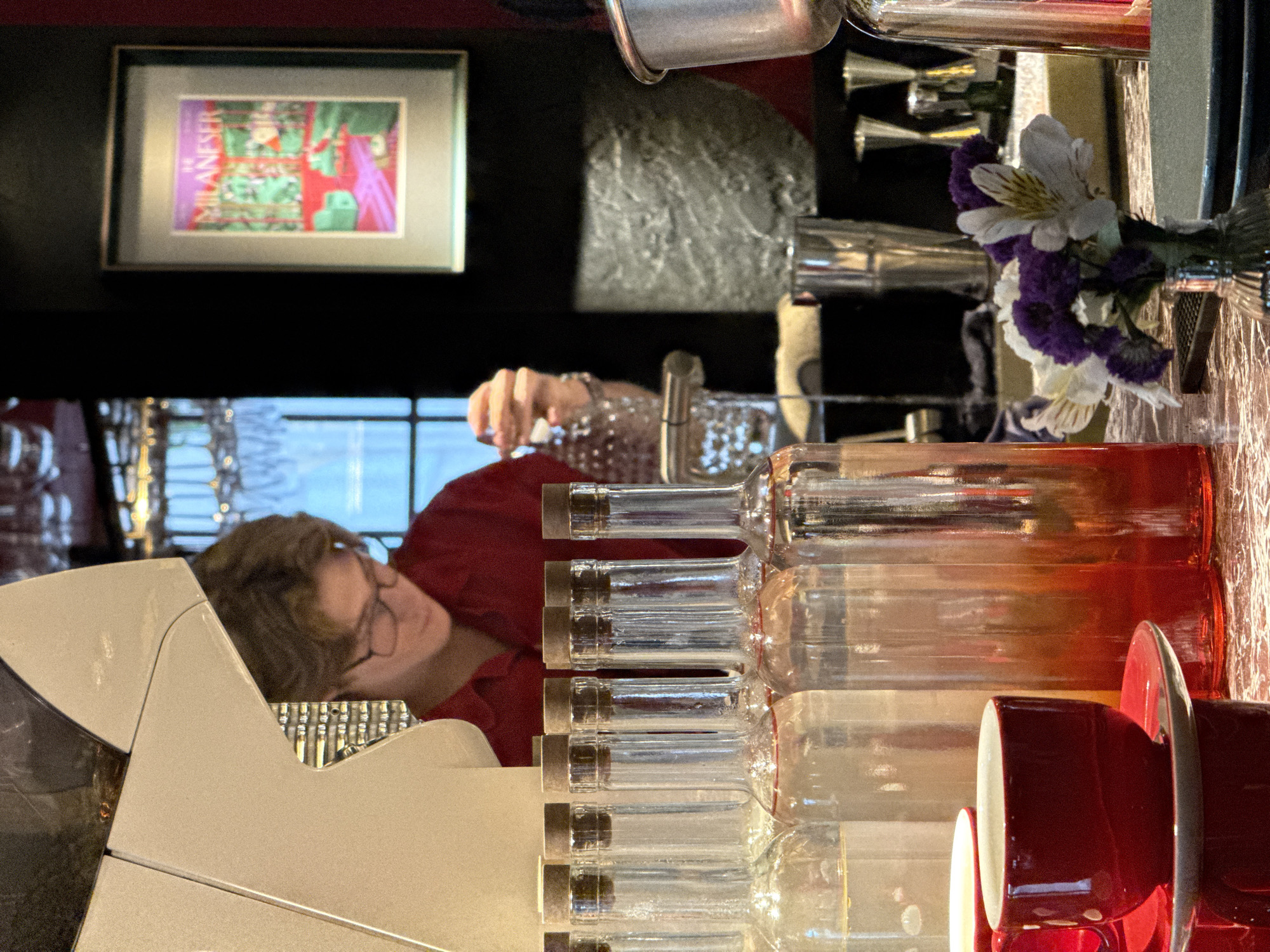 A person wearing glasses and a red shirt stands behind a bar, preparing drinks. Several glass bottles with cork stoppers are lined up on the bar. A framed artwork hangs on the wall in the background. In the foreground, a red coffee cup and saucer are visible. The scene appears to be inside a cafe or bar.