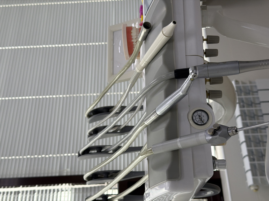 A dental station features a light gray control panel with various attachments. From top to bottom, there's a suction device, a high-speed handpiece, a low-speed handpiece, and a syringe with a nozzle. Each tool is connected by a clear hose to the central unit. A pressure gauge is visible, along with several adjustment knobs. A model of a set of teeth is mounted to the side, with a gray louvered wall in the background.