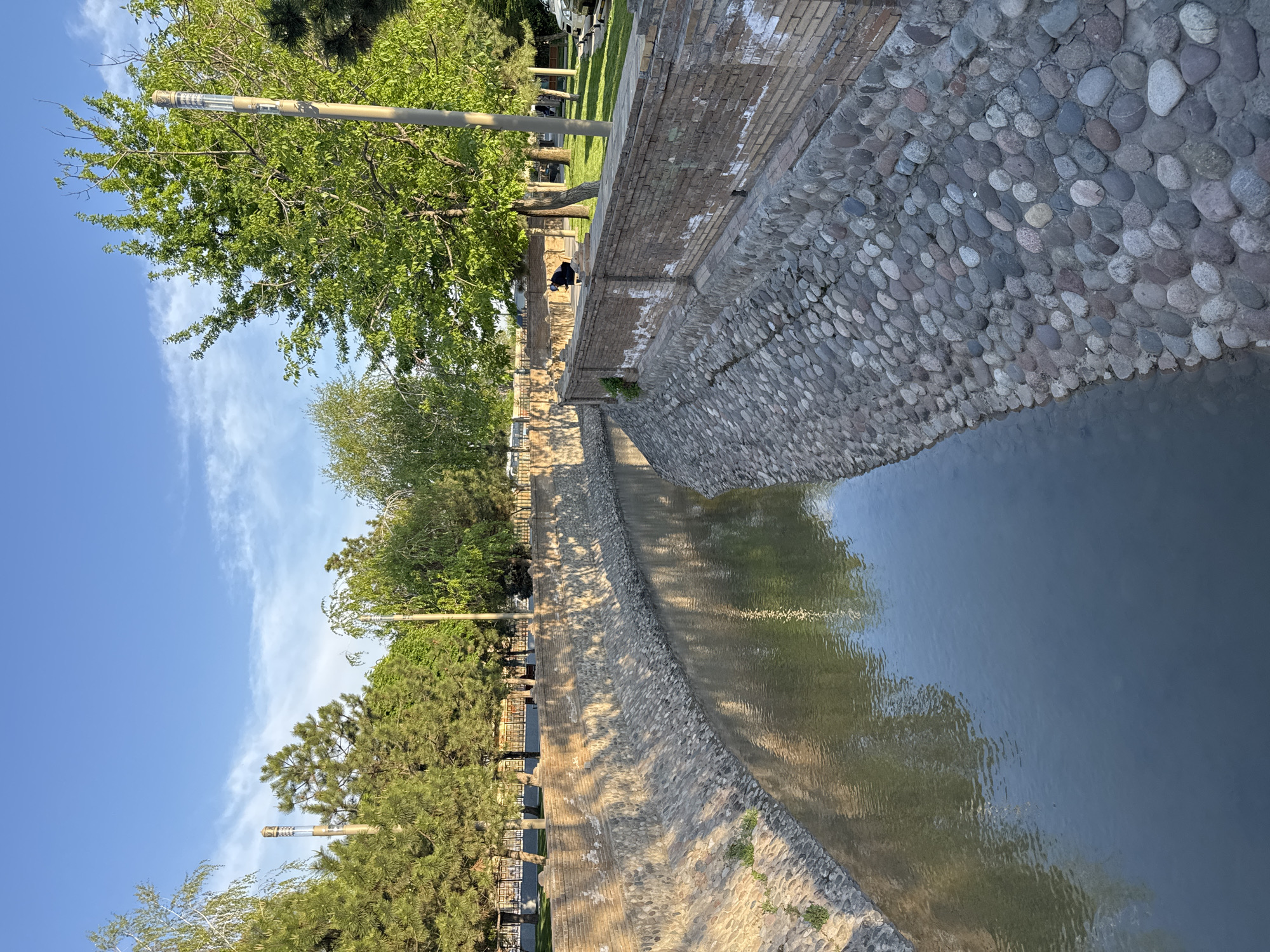 A water canal with stone embankments runs through a park in Tashkent, Uzbekistan. The embankment on the right is constructed of rounded cobblestones, while the embankment on the left is composed of stacked stone blocks. The canal water reflects the surrounding trees and sky. Trees line the canal, and a clear blue sky with scattered clouds is visible above. A person is visible sitting on the canal's edge in the middle ground. Street lights are seen lining the canal.