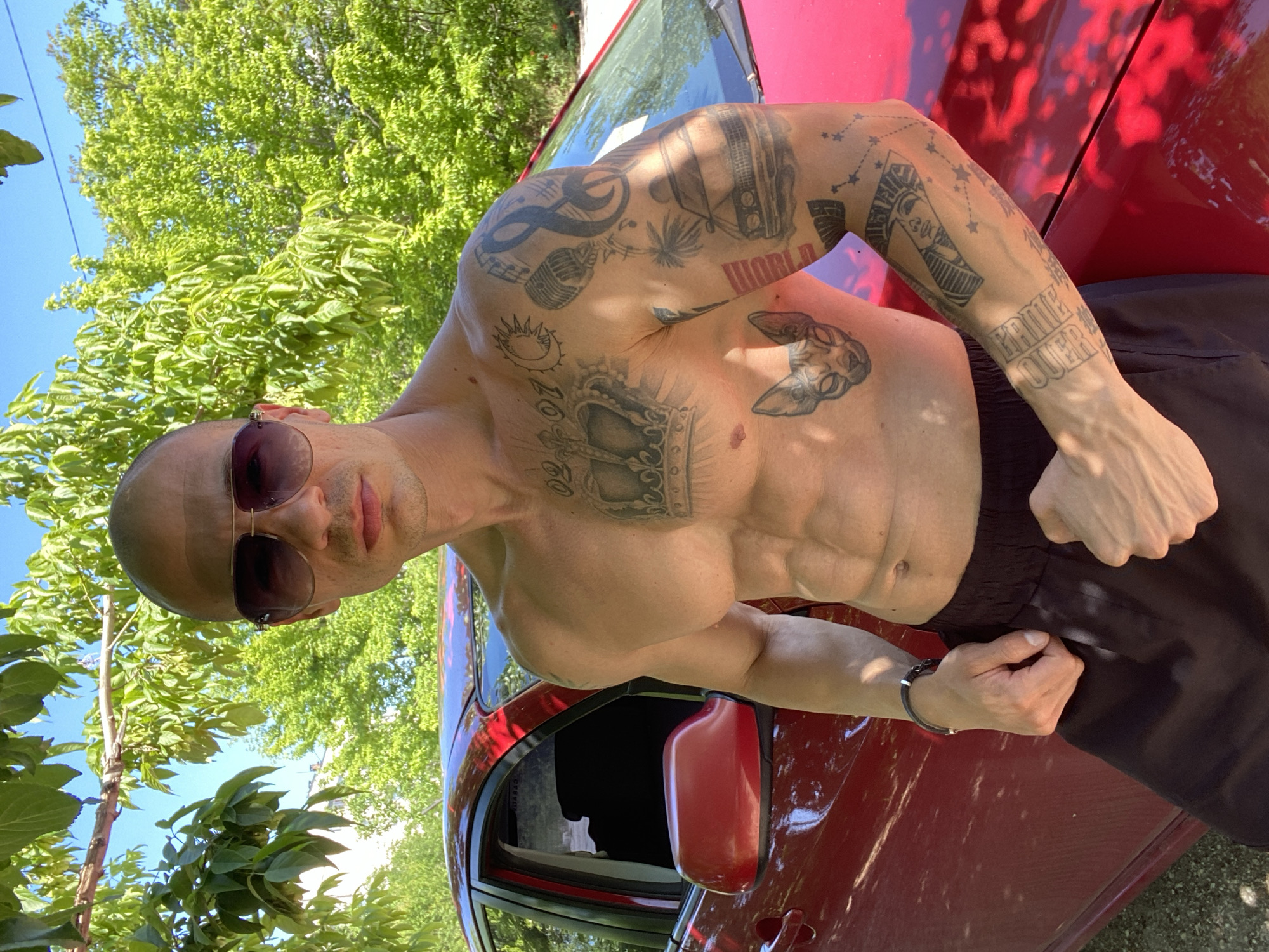 A bald, fair-skinned man with sunglasses stands shirtless next to a red car. He has numerous tattoos on his torso and arms. He wears dark-colored shorts. The background shows green trees and a blue sky, suggesting an outdoor location. It is not possible to determine the location in Pleven, Bulgaria from the image.