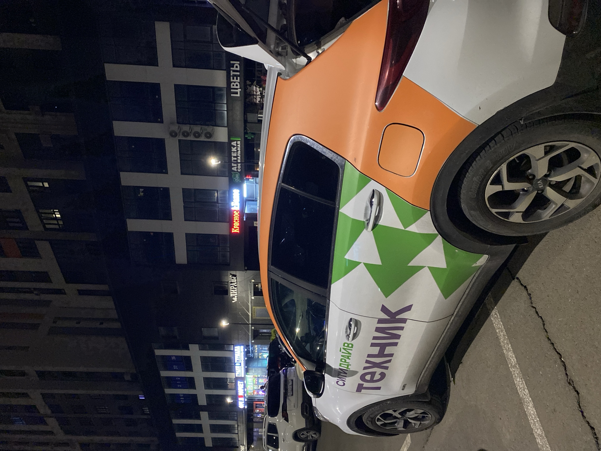 The image, captured at night, depicts a distinctive car parked on asphalt with tall, modern buildings in the background. The car, a white hatchback or SUV, features a vibrant livery with an orange rear half and a white front and mid-section adorned with a green and white geometric pattern. Large purple Cyrillic text on its side reads "СИТИДРАЙВ" (CityDrive) and "ТЕХНИК" (Technician), suggesting it belongs to a car-sharing or technical service. The vehicle's trunk/hatchback is open. Its rear right wheel, with a "KIA" logo on the hubcap and "Comfort 2 SUV" visible on the tire, is prominent in the foreground.
In the background, illuminated by streetlights and internal lights, stand several dark, multi-story buildings characterized by a grid-like facade and numerous windows. Various commercial signs, written in Cyrillic, are visible on these buildings: "ЦВЕТЫ" (Flowers), "АПТЕКА СОЦИАЛЬНАЯ" (Social Pharmacy), "ЛИРА" (Lyre/Lira), "ГРАНИ ГУРМАНА" (Gourmet Edges/Facets), and "Красное Белое" (Red White). Another white car is partially visible parked behind the main vehicle, displaying a license plate "E020V 797". The ground consists of asphalt with visible cracks and faint parking lines. The scene conveys an urban or commercial environment after dark.