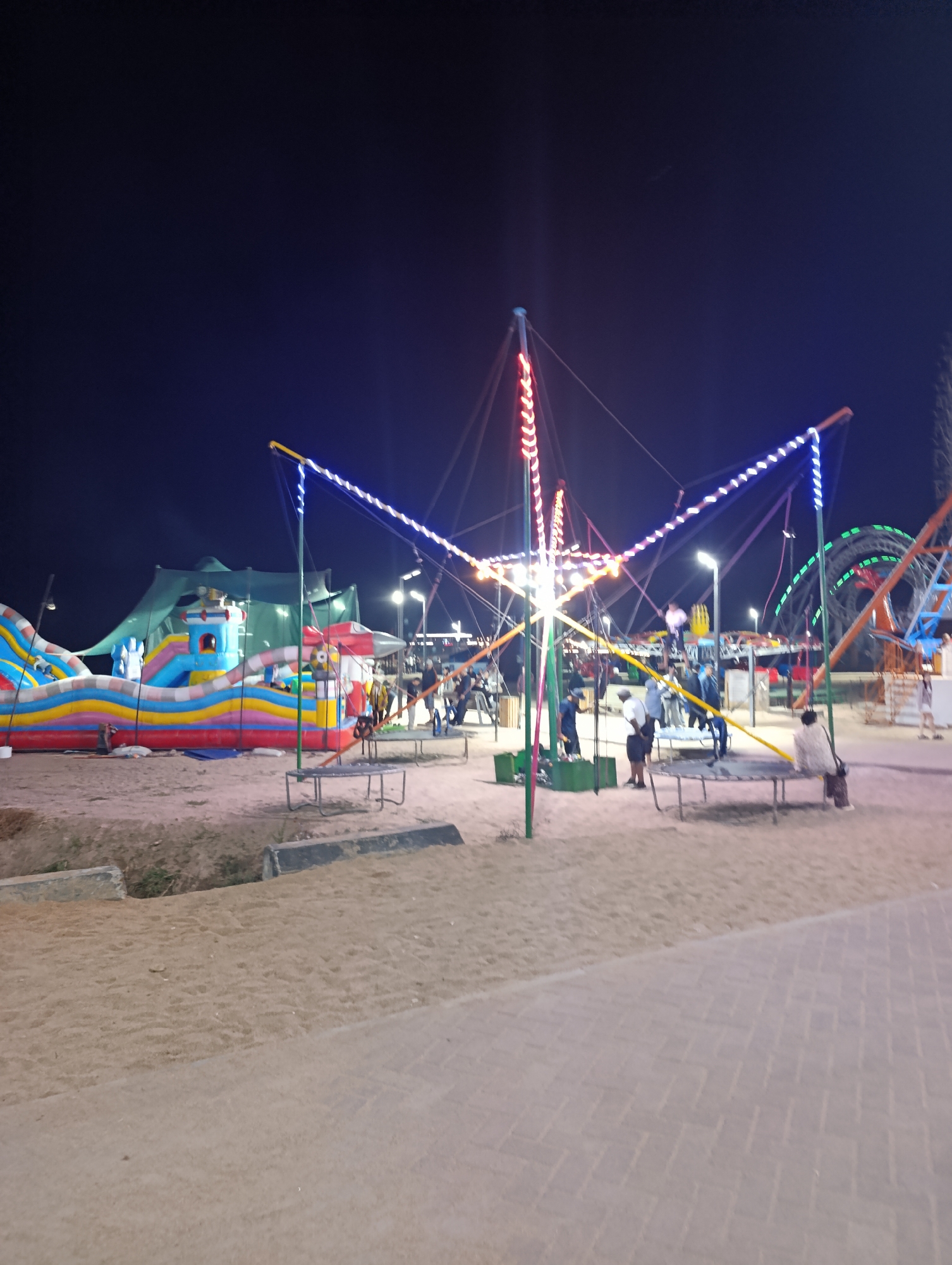 This image depicts a nighttime scene at an amusement park, possibly located in the city of Bosteri, Kyrgyzstan. In the foreground, a sandy ground leads to a paved walkway. The central focus is a bungee trampoline attraction with brightly colored LED lights. Several people are visible around this attraction, with some appearing to be waiting to use it. To the left, a large, inflatable bouncy castle with multiple slides is illuminated. In the background, the silhouette of a Ferris wheel or similar amusement ride can be seen against the dark night sky. The overall environment suggests a recreational area in an outdoor setting at night.