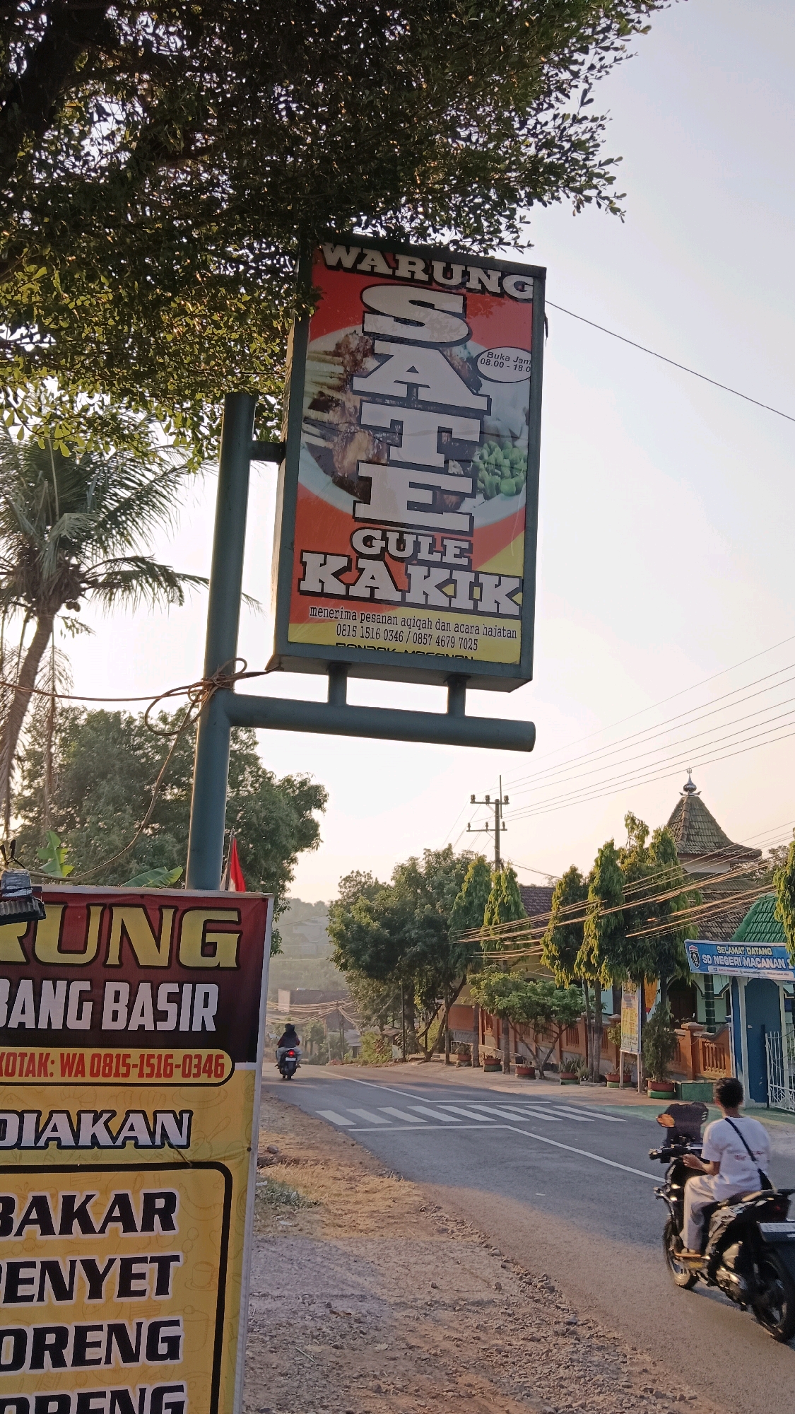 The image depicts a roadside scene in Jogorogo, Indonesia, at sunset. A prominent orange sign advertising "WARUNG SATE GULE KAKIK" (a type of satay and goat curry establishment) is positioned on the left, partially obscured by a tree. The sign also lists phone numbers and mentions "menerima pesanan aqiqah dan acara hajatan" (accepting orders for aqiqah and celebration events). Below this, another sign in yellow and brown is partially visible, with text including "BANG BASIR" and "KOTAK: WA 0815-1516-0346". Further down, "DI " and "BAKAR", "DENYET", and "ORENG" are visible, suggesting other food offerings.
On the road, a person is riding a motorcycle towards the viewer, followed by another person on a motorcycle in the distance. The road is paved and has white lane markings. The environment is rural, with trees, vegetation, and buildings visible on either side of the road. One building on the right appears to be a school, with a sign reading "SELAMAT DATANG SD NEGERI MACANAN 1" (Welcome to Macanan 1 State Elementary School). The sky is a soft orange, indicating the sun is setting.