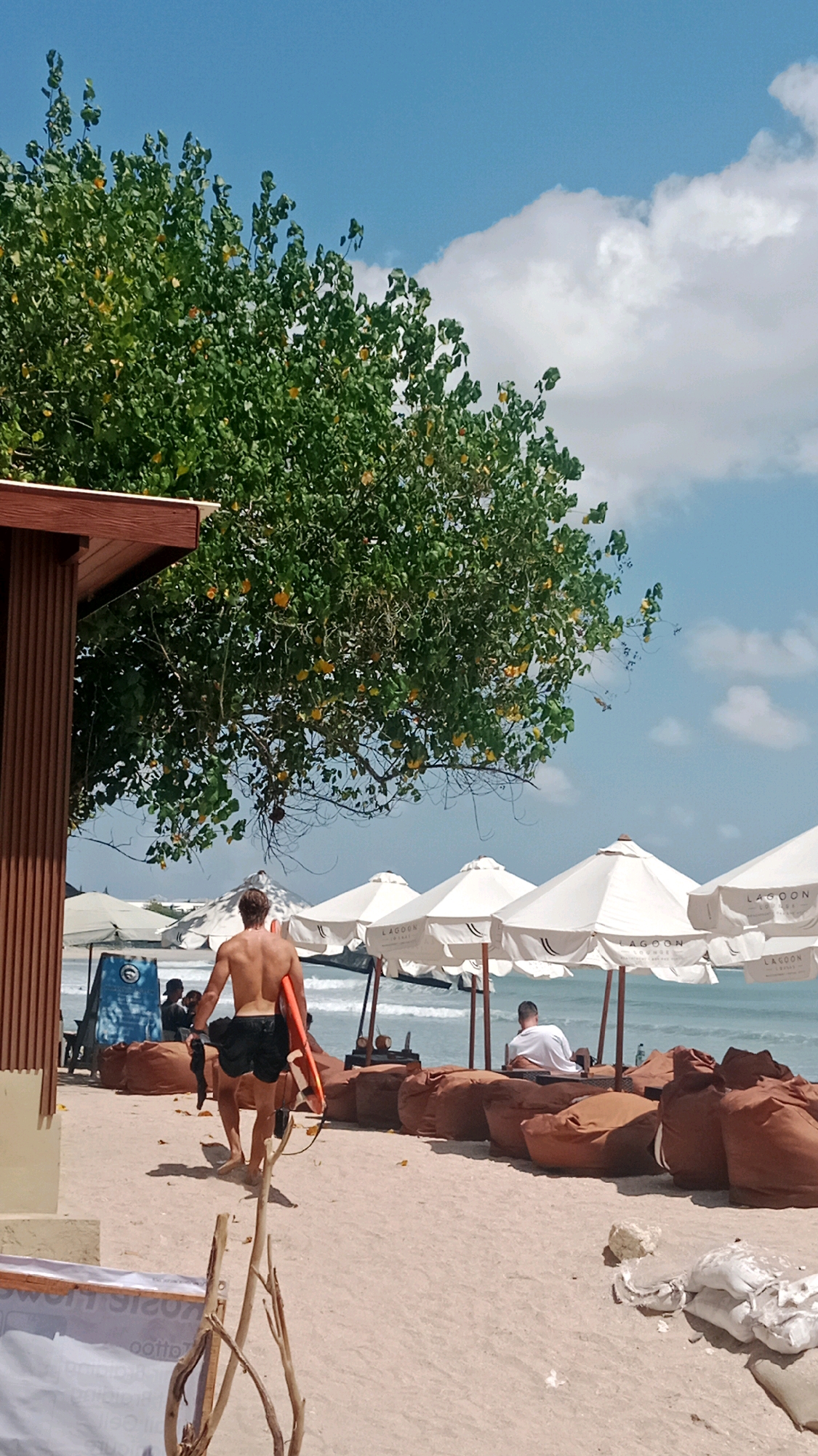 In Kuta, Indonesia, on a sandy beach under a bright blue sky with scattered white clouds, a young man with tanned skin and dark hair walks away from the camera, carrying an orange surfboard under his left arm. He is wearing black swim trunks and his back is to the viewer. To the left, a section of a wooden-sided structure with a brown roof is visible, partially obscured by a leafy green tree with small yellow fruits. In the background, several white beach umbrellas stand in a row on the sand, and the turquoise ocean meets the horizon. Numerous large, brown beanbag chairs are arranged in front of the umbrellas. Further back, other beachgoers are visible, some seated under umbrellas, and the waves of the ocean are gently rolling in. The scene suggests a relaxed atmosphere at a tropical beach resort.