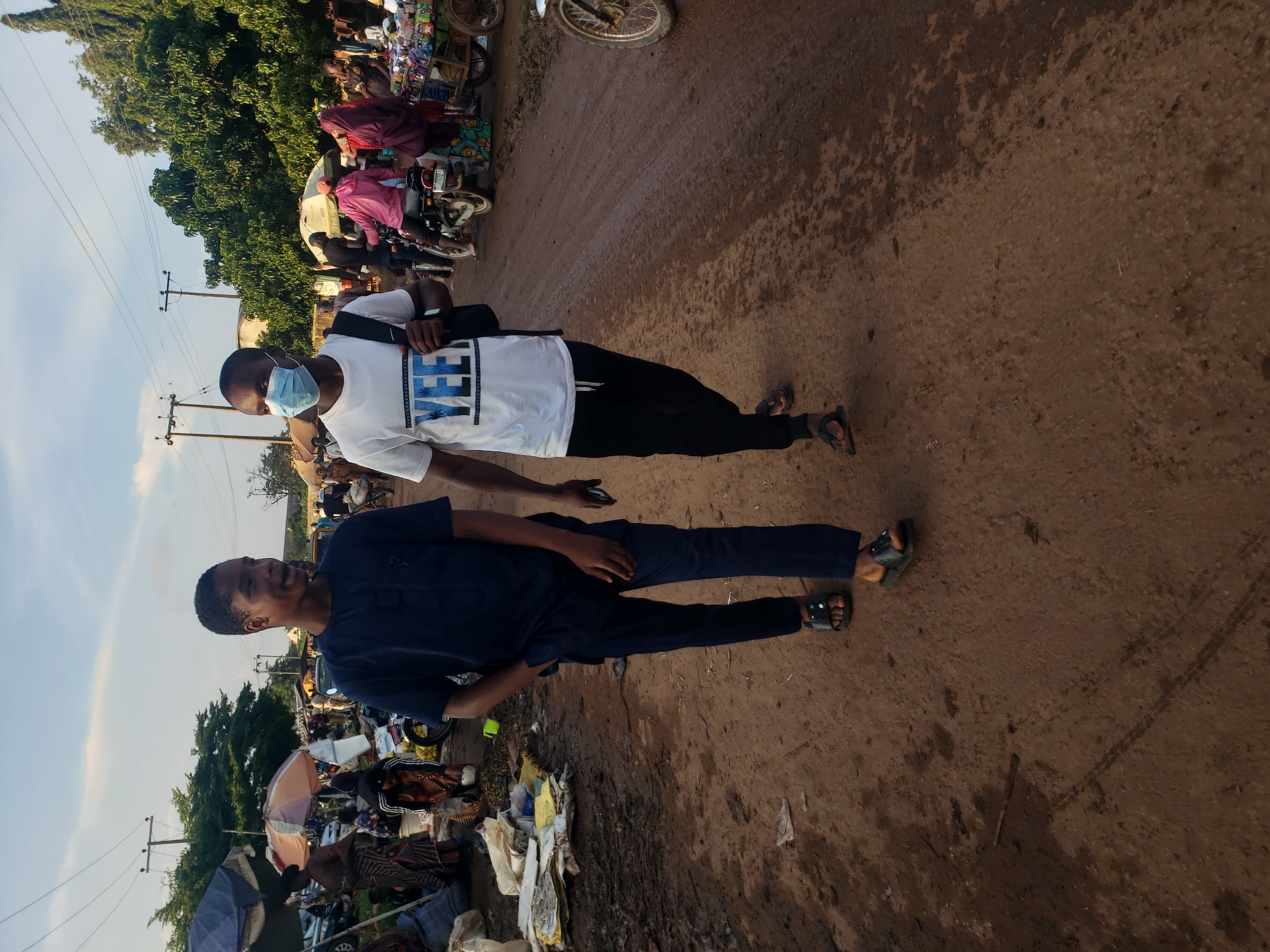 In Kakuri Gwari, Nigeria, two young men walk on a dirt road in an outdoor market. The man on the left wears a dark blue traditional outfit and sandals. The man on the right wears a white t-shirt with "YEET" printed on it in blue, black pants, and sandals, and a light blue face mask. He carries a black backpack over his right shoulder. The ground is muddy and dusty, with tire tracks visible. In the background, several market stalls are set up under umbrellas, with various goods displayed, and people are browsing. Motorcycles are parked and in use. Trees and power lines are visible in the distance under a partly cloudy sky.