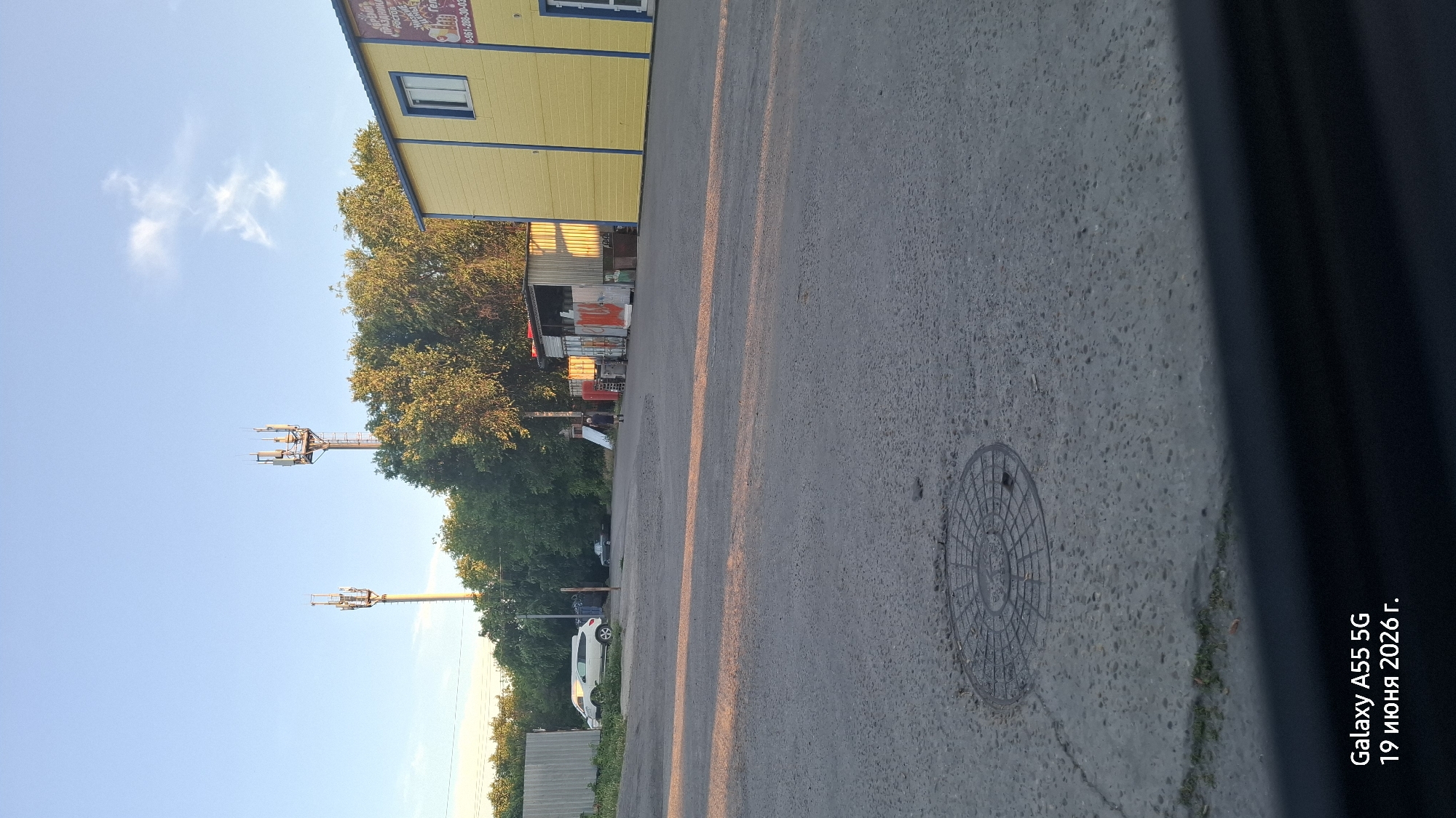 The image shows an outdoor scene in Rostov-on-Don, Russia, on June 19, 2026. In the foreground, a grey asphalt surface is visible with a manhole cover. The middle ground features a road leading towards a yellow building on the right, which has a sign with Russian text and a phone number. To the left of the yellow building, there are several sheds and containers. Trees line the background, and two telecommunication towers are prominent against a light blue sky with some clouds. A white SUV and a dark-colored car are parked near a metal fence on the left. A person is walking away from the camera towards the sheds on the left side of the yellow building. The lighting suggests it is late afternoon or early evening, with long shadows cast across the asphalt.