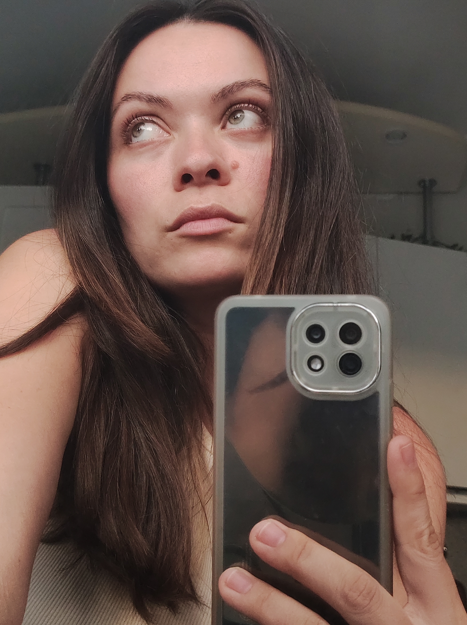 A woman is taking a selfie with her smartphone. She is looking off to the left, away from the camera. Her hair is long and brown. She has light skin, and her eyes are green. The smartphone has a clear case and a quad-camera setup. The background is indistinct, but a light fixture is visible. The location is not identifiable as Chaikovsky, Russia, based solely on the visual information provided in the image.