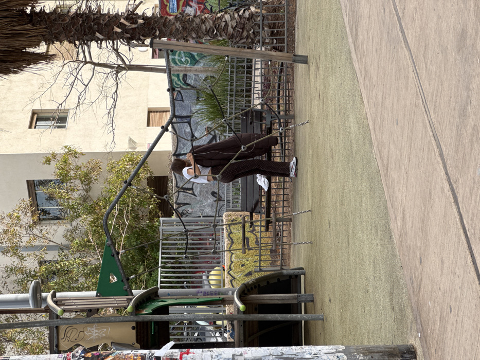 A person in a brown coat and patterned pants interacts with a rope structure in an outdoor area. The scene includes a building with windows, a palm tree overhead, and playground equipment. A paved path borders a grassy area. The background features graffiti art.