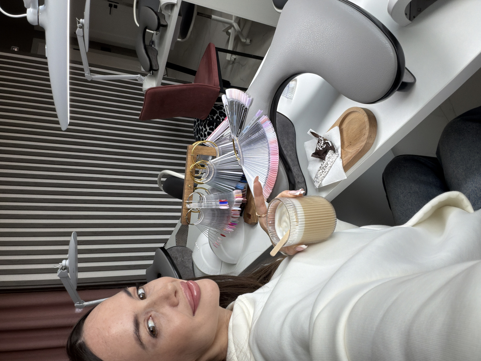 This selfie-style photograph captures a young woman in what appears to be a modern beauty salon or nail studio in Izhevsk, Russia. The image is rotated 90 degrees clockwise.
The central subject is a woman with long dark hair, brown eyes, and light pink lipstick, looking directly at the camera with a subtle smile. She is dressed in a white long-sleeved top or sweater. Her nails exhibit a light pink or white ombré manicure. In her right hand, she holds a ribbed, clear glass containing a creamy beverage, likely a latte, accompanied by a small wooden stirring stick.
The setting suggests a professional beauty establishment. On a white tabletop in front of her are several clear plastic fan-shaped displays, showcasing a comprehensive collection of nail polish colors, including various shades of pink, purple, blue, metallics, and neutrals. These swatches are arranged on a wooden stand with gold-colored rings. Adjacent to these, a small wooden tray holds a white napkin with a partially visible Ferrero Rocher chocolate, identifiable by the text "ROCHER" on its wrapper. A black-handled mirror is also placed on the table.
In the background, elements of the salon environment are visible, including parts of grey padded salon chairs, a reddish-brown upholstered chair, and a white adjustable lamp or light fixture. The wall behind features a distinctive pattern of horizontal black and white stripes, possibly wallpaper or blinds. The lower right side of the image shows a person's legs clad in dark jeans, suggesting another individual is seated nearby or the woman's own legs.
The scene depicts the woman appearing relaxed, likely waiting for or receiving a nail service, enjoying a drink and snack while potentially selecting her nail color. The overall ambiance is clean and modern.