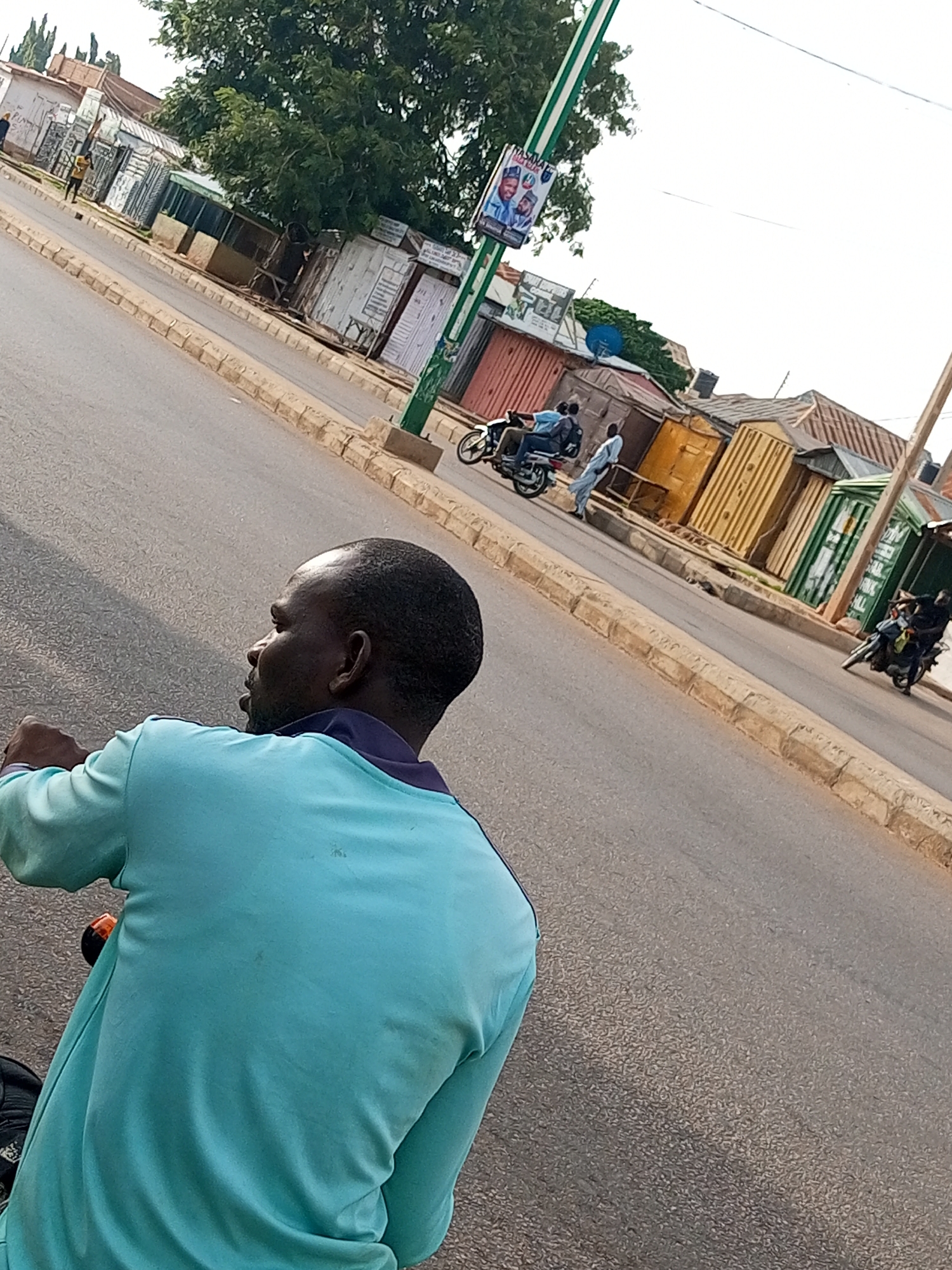 The image depicts a street scene in Gombe, Nigeria. In the foreground, a man is seen from the back, seated on a motorcycle. He is wearing a light blue long-sleeved shirt with a darker blue collar. His head is turned to the left, and his facial features are visible in profile. The motorcycle appears to be parked on the asphalt road.
The background of the image reveals a row of small shops or market stalls, constructed from what appear to be corrugated metal and other salvaged materials. Some of these structures are painted in muted colors, while others, like a bright yellow one, stand out. A large tree with dense foliage is visible behind some of the stalls, providing shade. A green pole with a poster attached is also present. Further down the street, two other motorcycles with riders are visible, and a person in a light-colored garment is walking. The road is lined with a raised curb made of beige-colored stones. The sky is overcast, suggesting it might be a cloudy day.