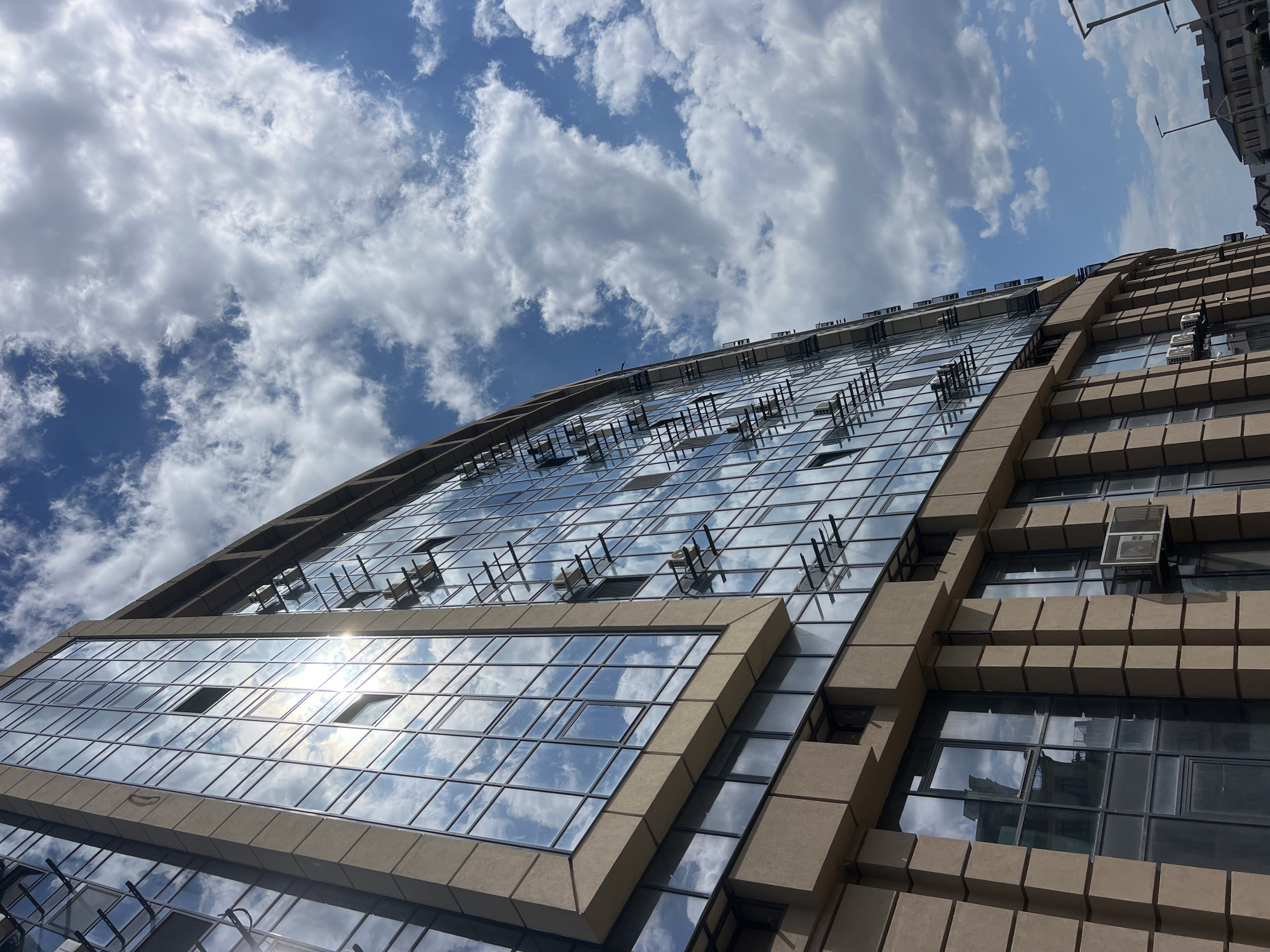 The image shows a modern building with a facade primarily composed of glass windows and beige stone panels. The windows reflect the blue sky and scattered white clouds. Some windows are open, revealing the interior structure. Air conditioning units are visible mounted on the exterior walls. The building appears to be in an urban environment, with streetlights and other buildings visible in the background. The perspective is from a low angle, looking up at the building against the sky. The location is identified as Fontanka, Ukraine.