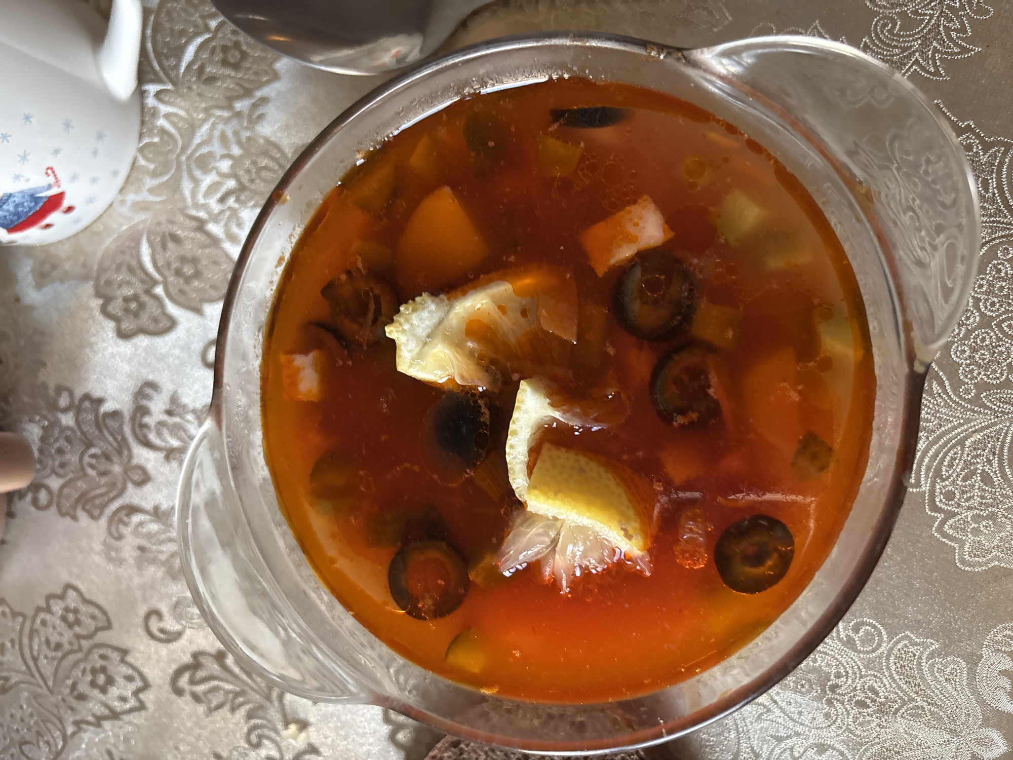 A close-up, overhead shot showcases a clear glass bowl filled with a reddish-orange soup. The soup contains visible ingredients such as sliced black olives, what appear to be small pieces of white cheese or possibly seafood, and wedges of lemon. The bowl rests on a patterned tablecloth with a floral design in beige and white tones. A partial view of a mug with a festive design, possibly a Christmas theme, is visible in the upper right quadrant of the image, along with a hint of a person's hand in the upper left. A silver spoon or ladle is partially visible on the right edge of the frame. The scene suggests a meal setting, possibly in a home environment. The exact location in the city of Unknown, Russia is not discernible from the image alone.