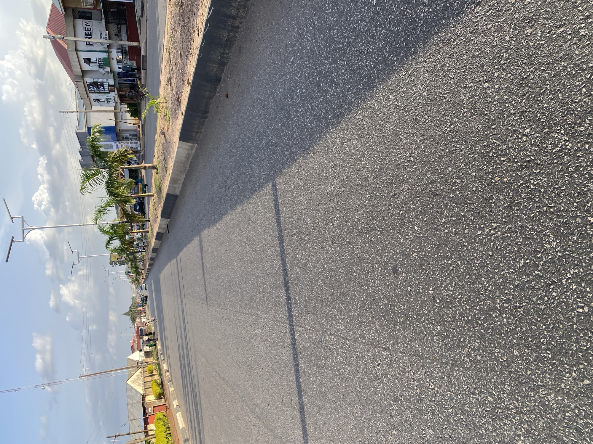 This image captures a street scene in Jos, Nigeria, characterized by a paved road with a distinct asphalt texture. The road is bordered by a concrete curb and a sandy shoulder, with sparse vegetation and a few palm trees lining the side. Buildings, some appearing commercial with storefronts, are visible in the background, including one with "FREE TV" signage. Several people are present, including some walking on the sidewalk and others near the buildings. Vehicles, including cars and what appears to be a tricycle taxi, are on the road. Overhead, a utility pole with antennas and streetlights are visible, indicating urban infrastructure. The sky is partly cloudy, suggesting a daytime scene with clear weather conditions. The overall impression is of a typical urban street in Jos.