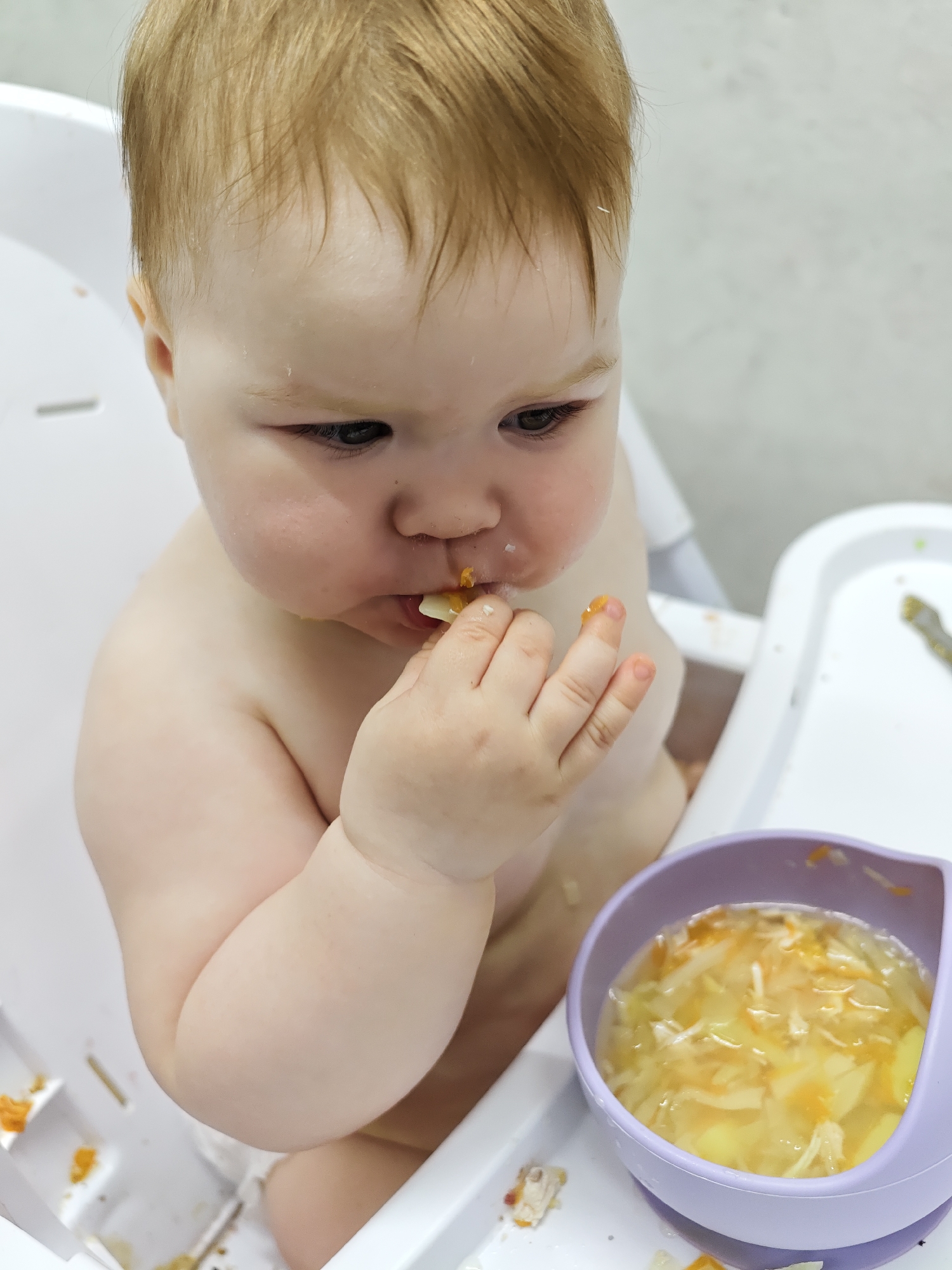 The image depicts a baby sitting in a highchair, apparently eating food with their hands. The baby has fair skin, light brown hair, and is wearing what appears to be a diaper. The baby is holding a piece of food near their mouth, and there is food residue on their face and fingers. To the right of the baby is a purple bowl containing a broth-based soup with visible vegetables. The highchair is white and is placed against a light-colored wall. The location is unspecified, and there is no indication it is in the city of Chaikovsky, Russia.