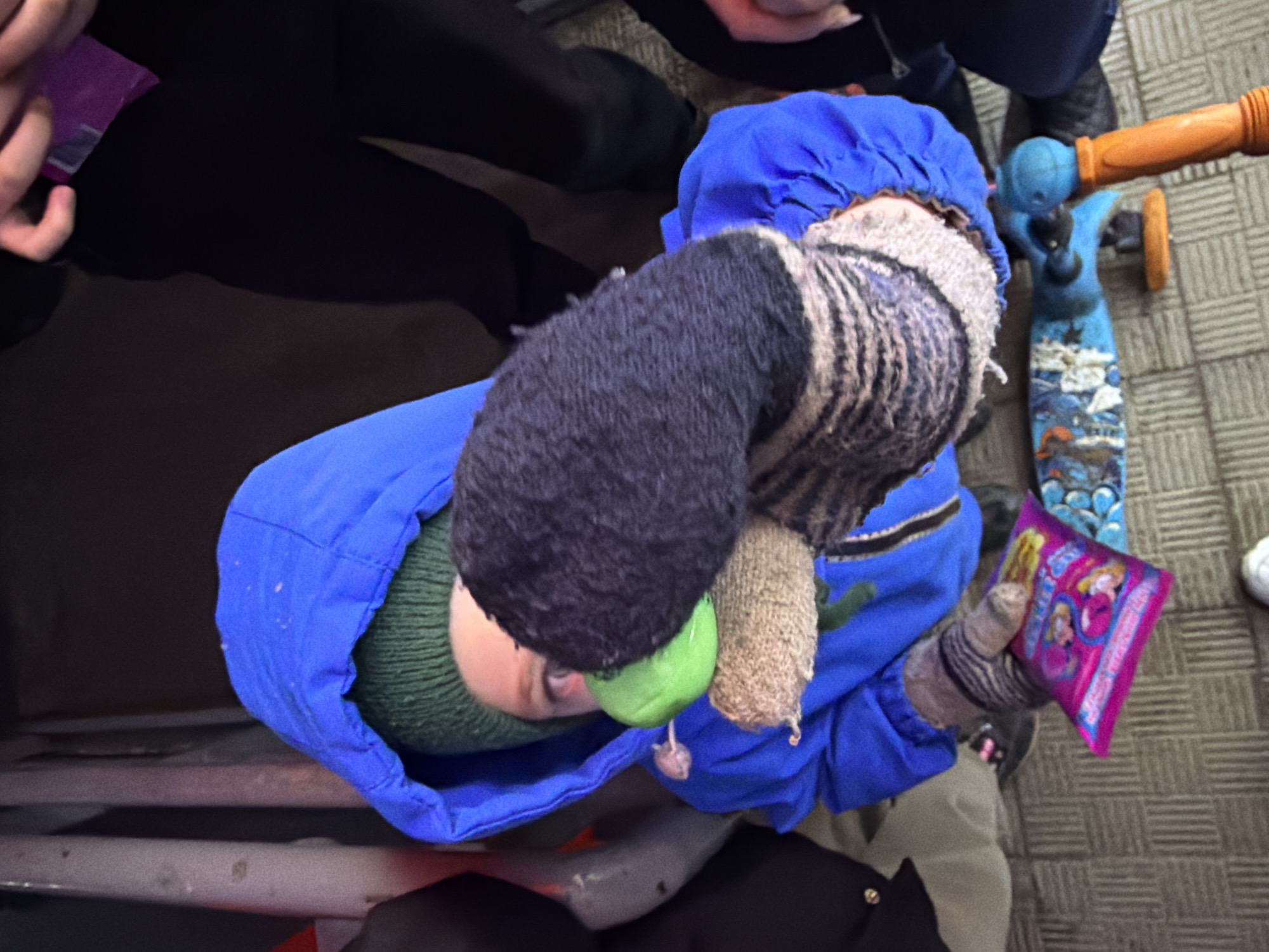 An overhead shot captures a young child looking upwards, dressed in a bright blue hooded jacket, a green knitted hat, and grey and white striped gloves. Attached to or held by the child is a long, multi-textured knitted item, featuring dark grey, light brown, and cream sections, from which a vibrant green, apple-shaped object dangles. In the child's gloved right hand, a small, rectangular, vibrant purple packet is held, displaying cartoon characters, likely princesses, and the word "Princess" is discernible among other indistinct English text.
To the right of the child, a blue and orange scooter with graphic designs on its deck rests on a light-colored, patterned floor. Partial views of an adult, wearing black trousers and shoes, are visible at the top and left of the frame. The scene appears to be set indoors, possibly within a public building or a home, characterized by the patterned floor tiles. The child seems engaged with their items and is looking up, suggesting interaction with someone or something above them. The overall ambiance is casual and everyday. No specific time of day or weather can be determined from the indoor setting. The location is Chaikovsky, Russia.