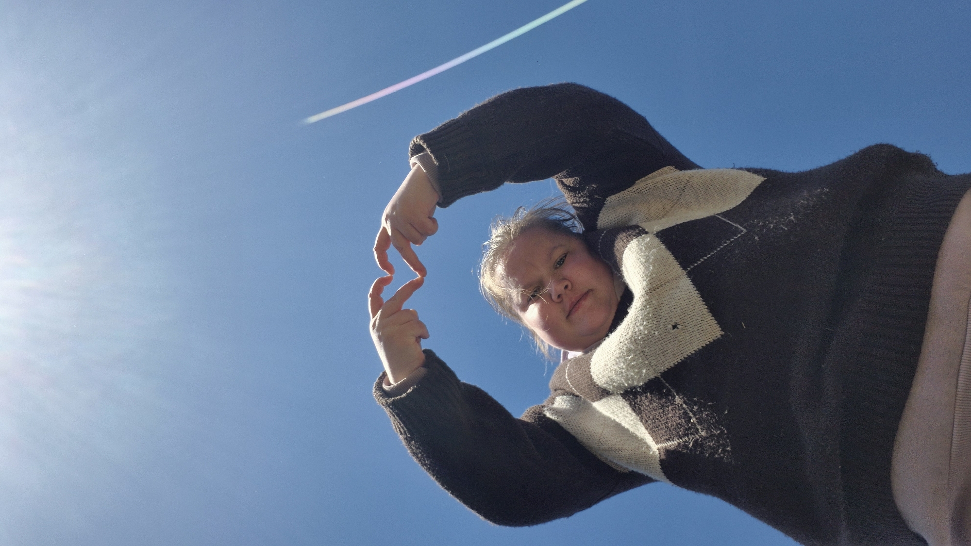 A low-angle shot against a clear blue sky captures a person holding their hands up in front of their face. The person is wearing a dark, patterned sweater with light-colored accents. A bright streak of light is visible in the upper left portion of the sky. The photograph does not contain information that can determine a specific location in Дойная, Russia.