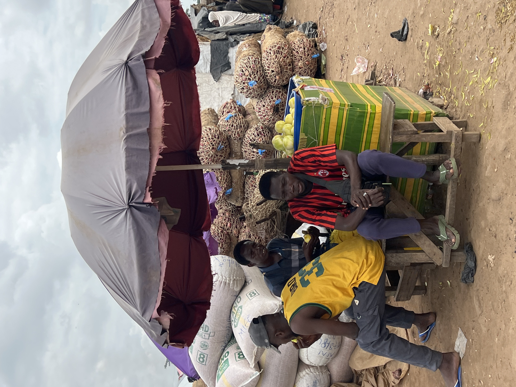 The image depicts an outdoor market scene in Oko Amakom, Nigeria, under a large, tattered umbrella providing shade. Three men are visible. The man on the left, wearing a yellow and green tank top and a grey baseball cap, is seated and appears to be eating a piece of fruit. In the center, a man in a blue checkered shirt is also seated and holding a piece of fruit. On the right, a man in a red and black striped t-shirt is seated, facing forward.
Behind the men, piles of goods are stacked. To the left are large white sacks, some with visible markings. To the right and center are numerous sacks or bundles of onions, contained within netting. A green and yellow striped cooler or display box is positioned in front of the man on the right, holding a pile of green, round fruits, possibly limes or small gourds.
The ground is composed of packed dirt and sand, with scattered debris. The sky is overcast, suggesting a cloudy day. The overall impression is that of a bustling, informal marketplace where vendors and customers are gathered.