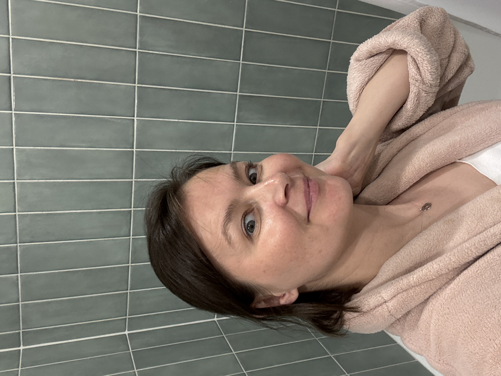 A woman is pictured in a bathroom. She is wearing a pink bathrobe, with the hood up and her arm inside. The bathroom has grey-green, horizontally-aligned subway tile on the walls. The woman is facing towards the camera, with her head tilted to the right. She has short brown hair, light skin, and is looking at the camera. There are no indications about the location in the city of Izhevsk, Russia.