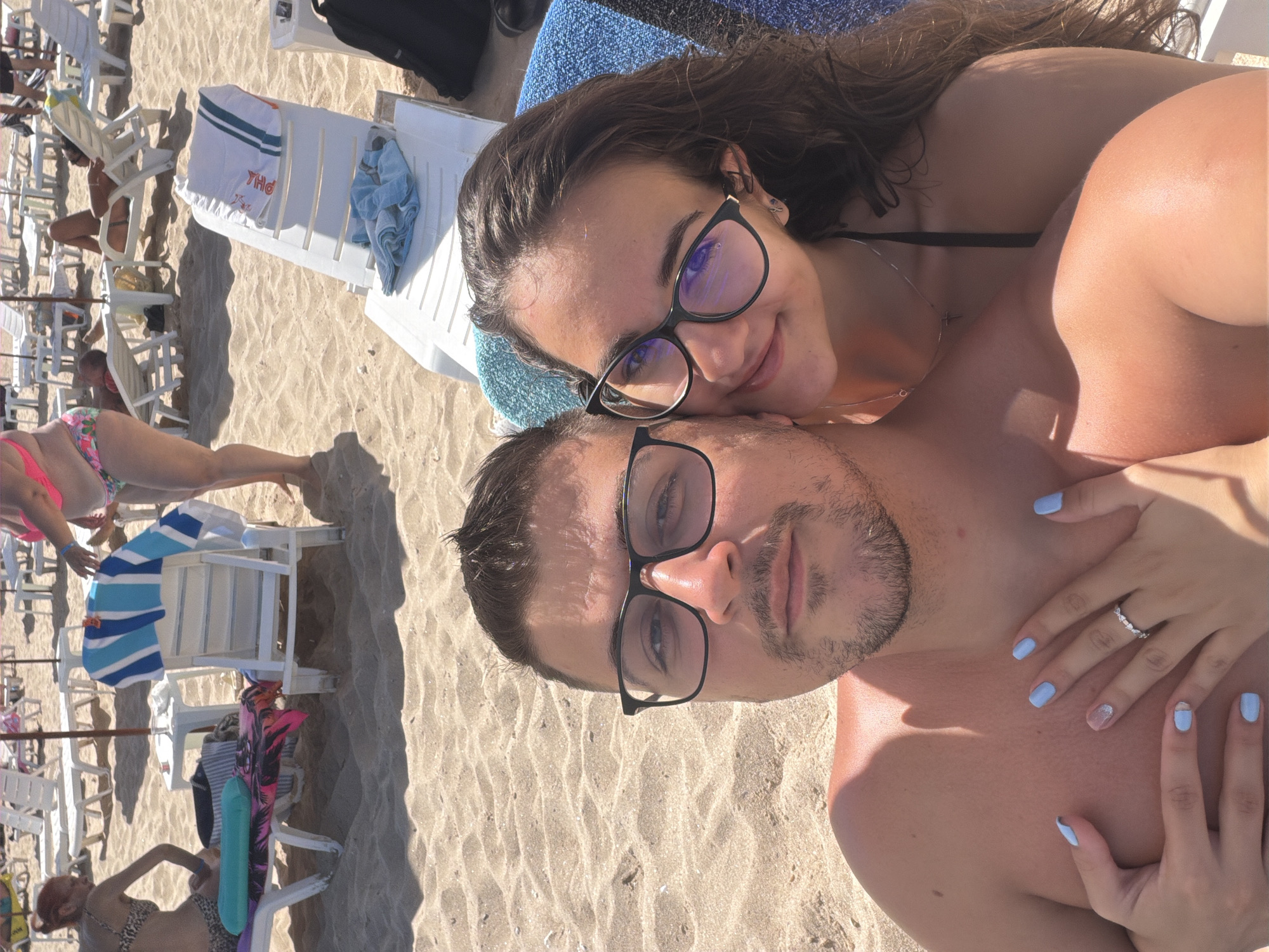 A couple, a man and a woman, are taking a selfie on a sandy beach in Primorsko, Bulgaria. The man, wearing glasses, is shirtless and has his arm around the woman, who is also wearing glasses and a black bikini top. They are both smiling at the camera. In the background, beach chairs and umbrellas are visible, and other beachgoers are present. The sand is light brown and appears to be warm and dry. The lighting suggests it is a sunny day.