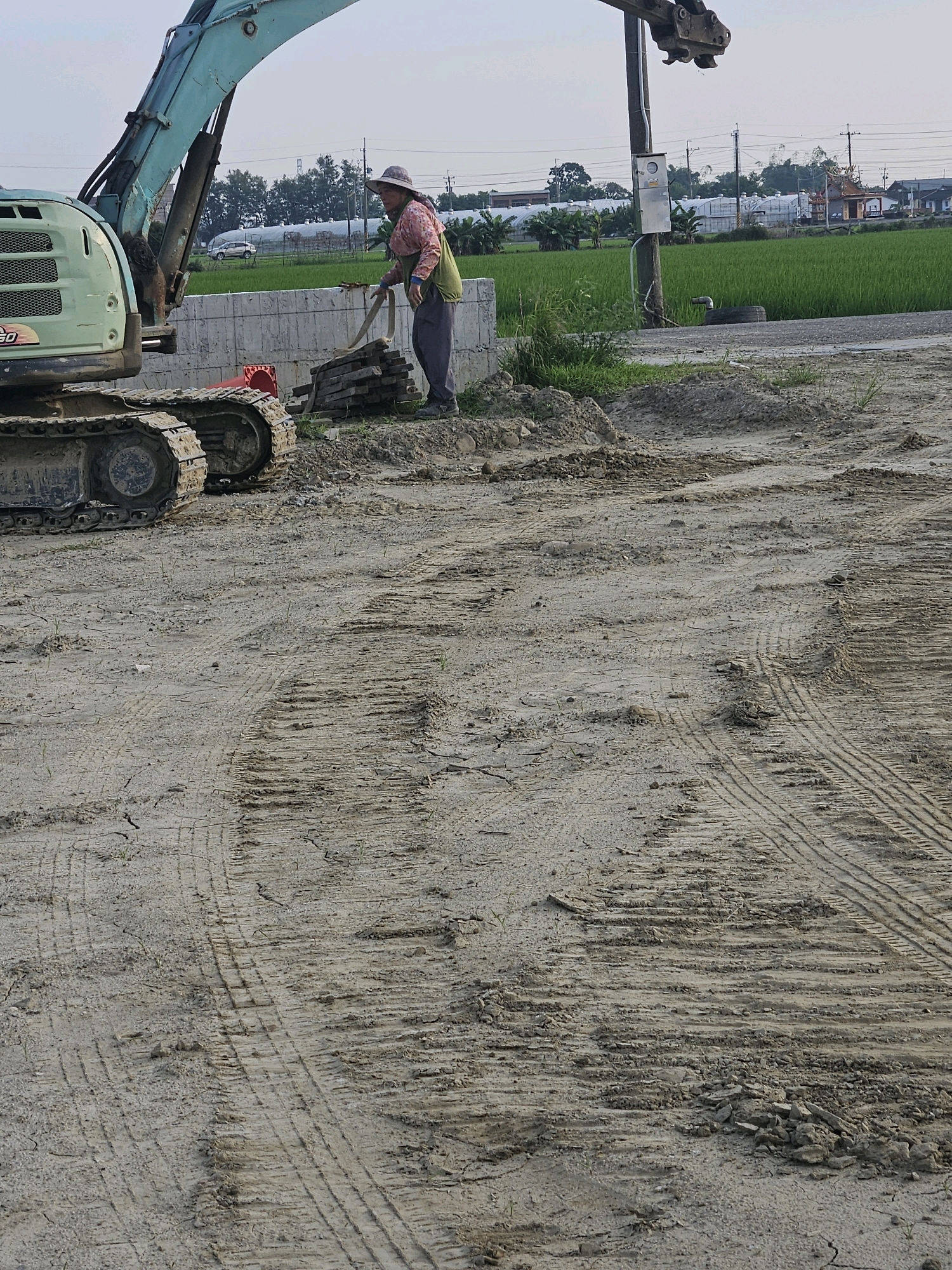 An excavator sits on a dirt construction site in Xikou Township, Taiwan. A person wearing a hat and a pink and green shirt is shoveling bricks near the excavator. In the background, there is a green field of rice, some greenhouses, power lines, and a small building with a red roof. The ground is dry and cracked, with visible tire tracks from heavy machinery. The sky is overcast.