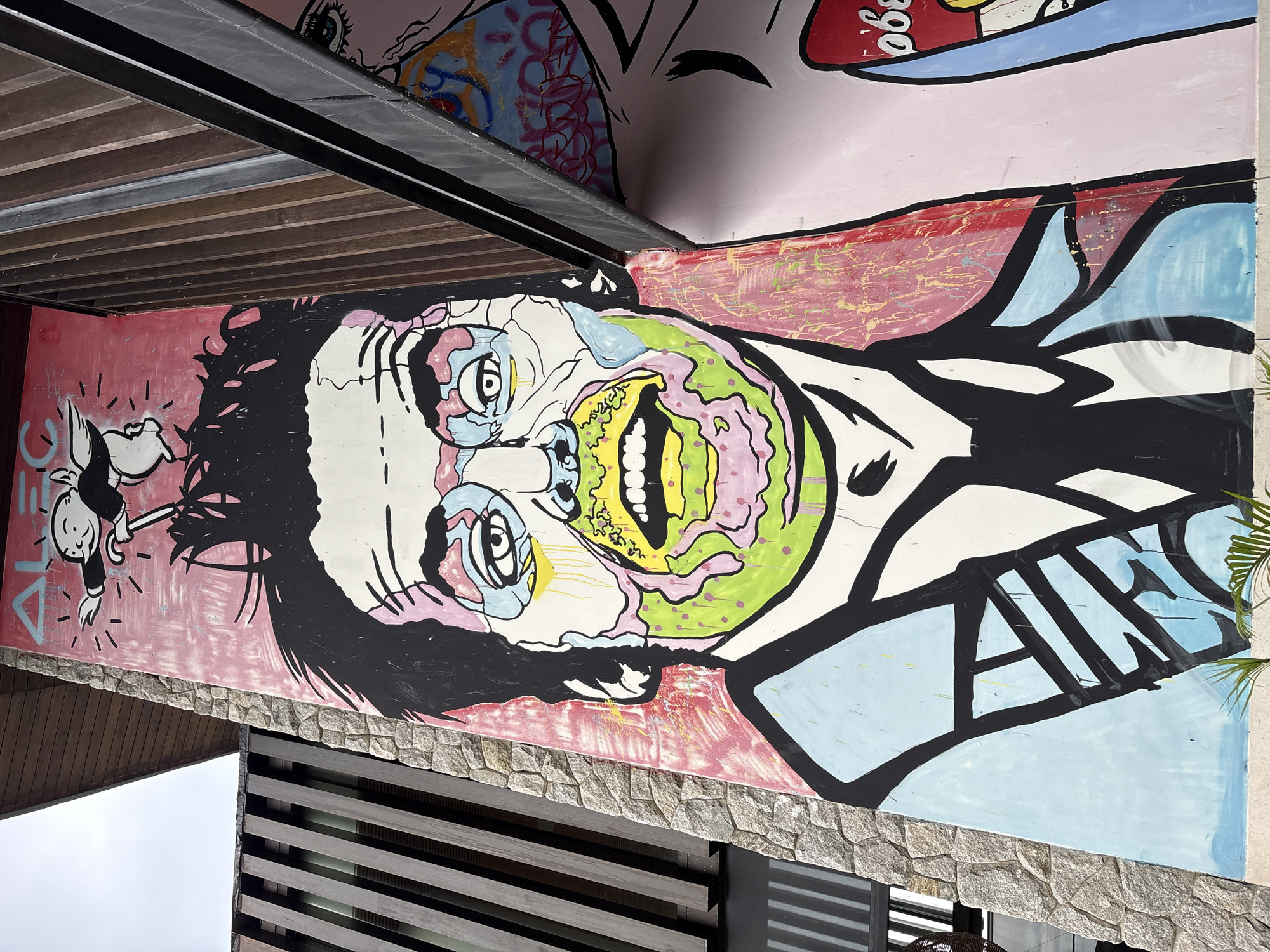 A vibrant mural dominates the scene in Baan Bo Phut, Thailand. The artwork features a grotesque yet captivating face with exaggerated features and psychedelic coloring. The face is predominantly white, with black hair and outlines. The eyes are a mix of blue and yellow, while the mouth is a vivid green and yellow, revealing a row of white teeth. Pink and green splashes decorate the cheeks and neck, which merges into a light blue shirt. To the left of the face, a smaller figure in a suit hangs suspended. The background is primarily a pinkish-red, transitioning to a light blue on the right. The mural is painted on a wall made of rough, light-colored stones. A modern building structure with wooden slats is visible below and beside the mural. Above the mural, structural supports and a partial view of another mural with graffiti-style elements are visible. A small portion of a green palm frond is seen on the right.