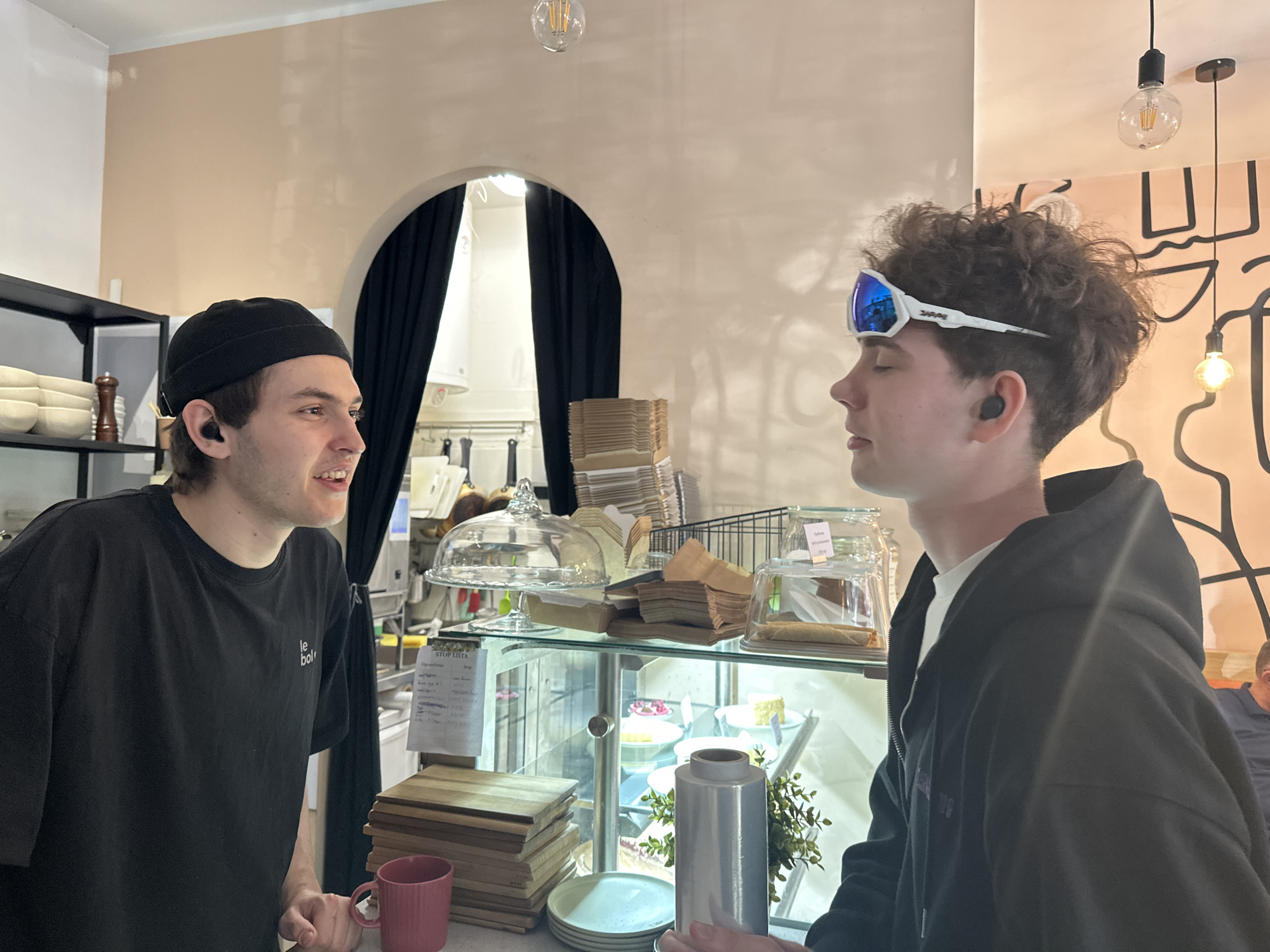 The photograph captures an interaction between two individuals in what appears to be a cafe or bakery setting. On the left, a young man with dark hair, wearing a black beanie and a black t-shirt with small text ("le bol") is speaking to another individual. He is standing behind a counter with a pink mug and stacks of wooden cutting boards.
The second person, standing on the right, is a young man with curly hair and white framed glasses with blue lenses resting on his forehead. He is wearing a black hooded jacket over a white t-shirt.
Behind them is a glass display case containing pastries, cakes, and other food items. Wrapped food, plates and a cylindrical metal container rest on the counter. Behind the display case, an arched opening is draped with heavy black curtains, suggesting entry to a kitchen. A light fixture is visible through the arch. Above the counter, additional pastries and baked goods are visible. The background features light fixtures and a stylized pattern on the wall.
The scene is brightly lit, highlighting the details of the cafe setting. The location is Novi Sad, Serbia.
