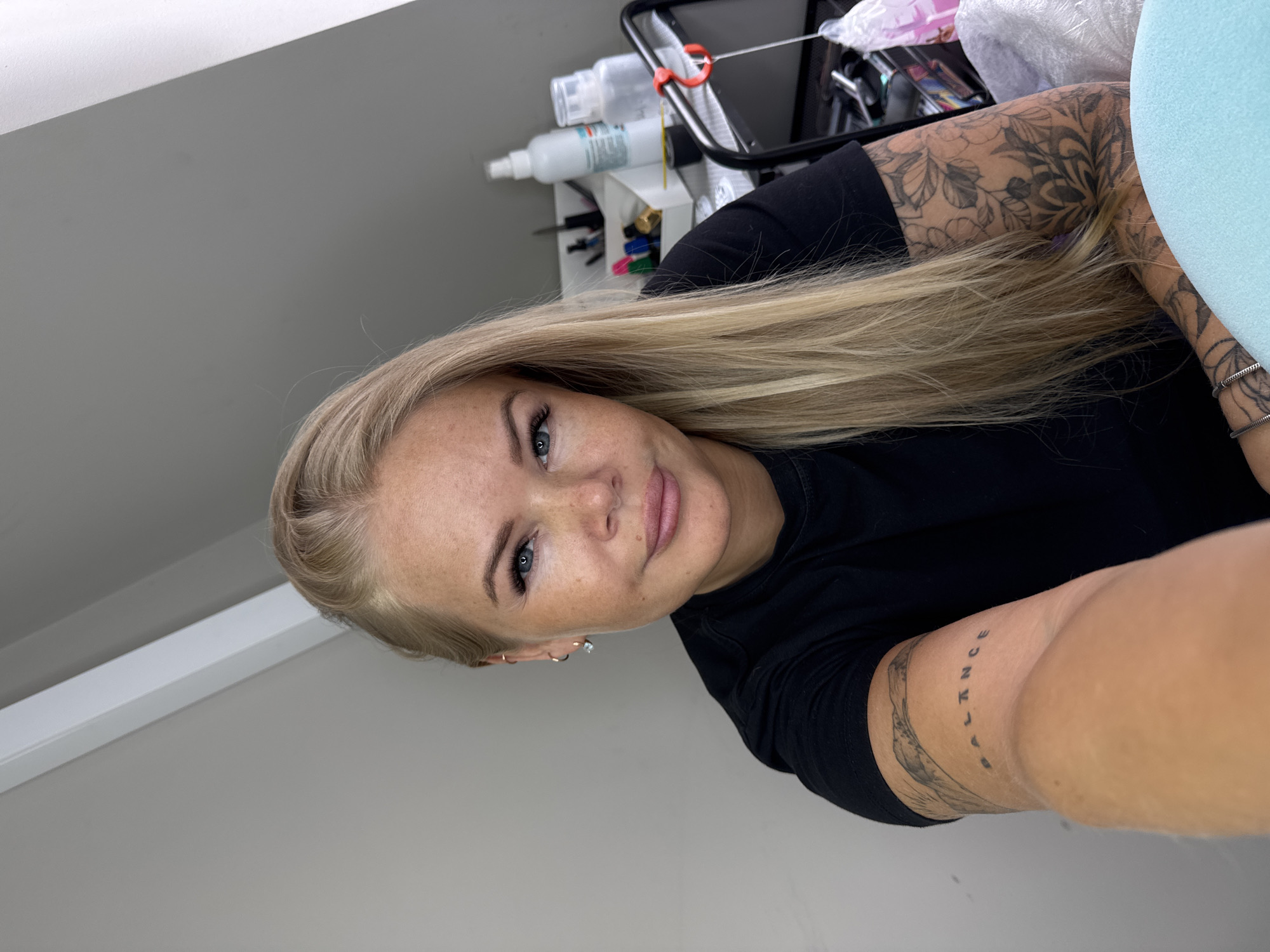 A fair-skinned woman with blonde hair is positioned in front of a light grey wall and white ceiling. She has a tattoo on her left inner forearm and arm. A cosmetic storage cart containing bottles, containers, and writing utensils is visible behind her left arm. She is wearing a black top and there is a large blue pillow to her right.