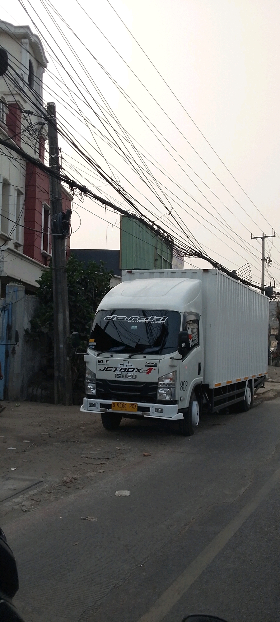 A white Isuzu Elf truck with a white container body is parked on a paved road. The truck has a license plate that reads "B 9184 PXX". The truck has decals on the windshield that say "daraki" and "BUJANG NYEN". The truck also has "ELF JETBOX" and "ISUZU" emblems on the front. The truck is parked in front of a building with red and white walls. The street is lined with power lines. The sky is overcast. The scene appears to be in an urban environment.