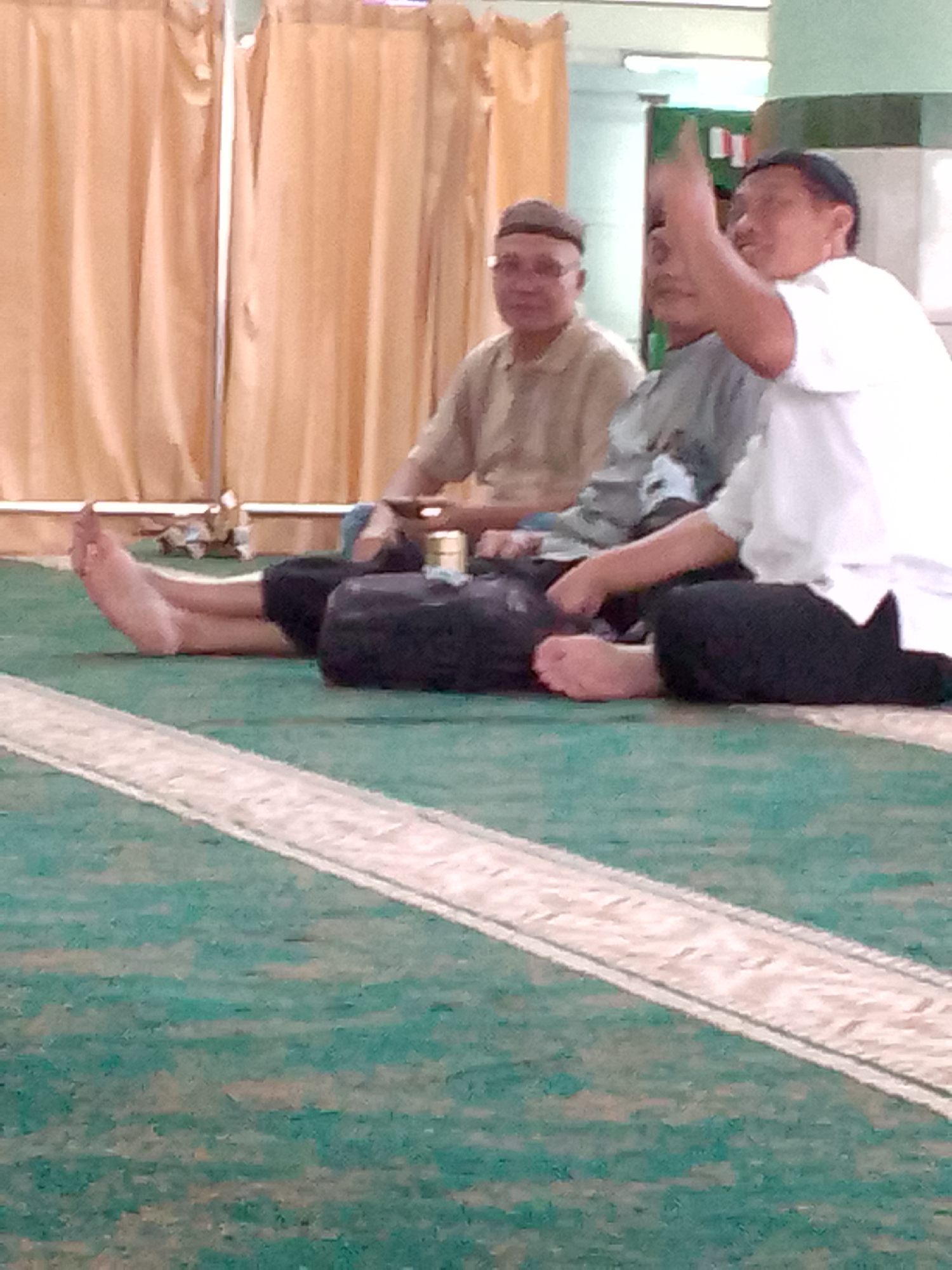 Three men are seated on a green carpet with a white stripe pattern, likely in a prayer hall or communal gathering space. The man on the far left wears glasses and a light-colored collared shirt with a cap. The man in the middle is wearing a grey shirt and has his face partially obscured. The man on the right, wearing a white shirt, is gesturing with his right hand, index finger raised, and looking towards the viewer. A dark bag rests on the floor in front of the two men on the left. In the background, peach-colored curtains hang, and a pale green pillar is visible. The image appears to be taken in Cimahi, Indonesia, possibly at a mosque or religious center.