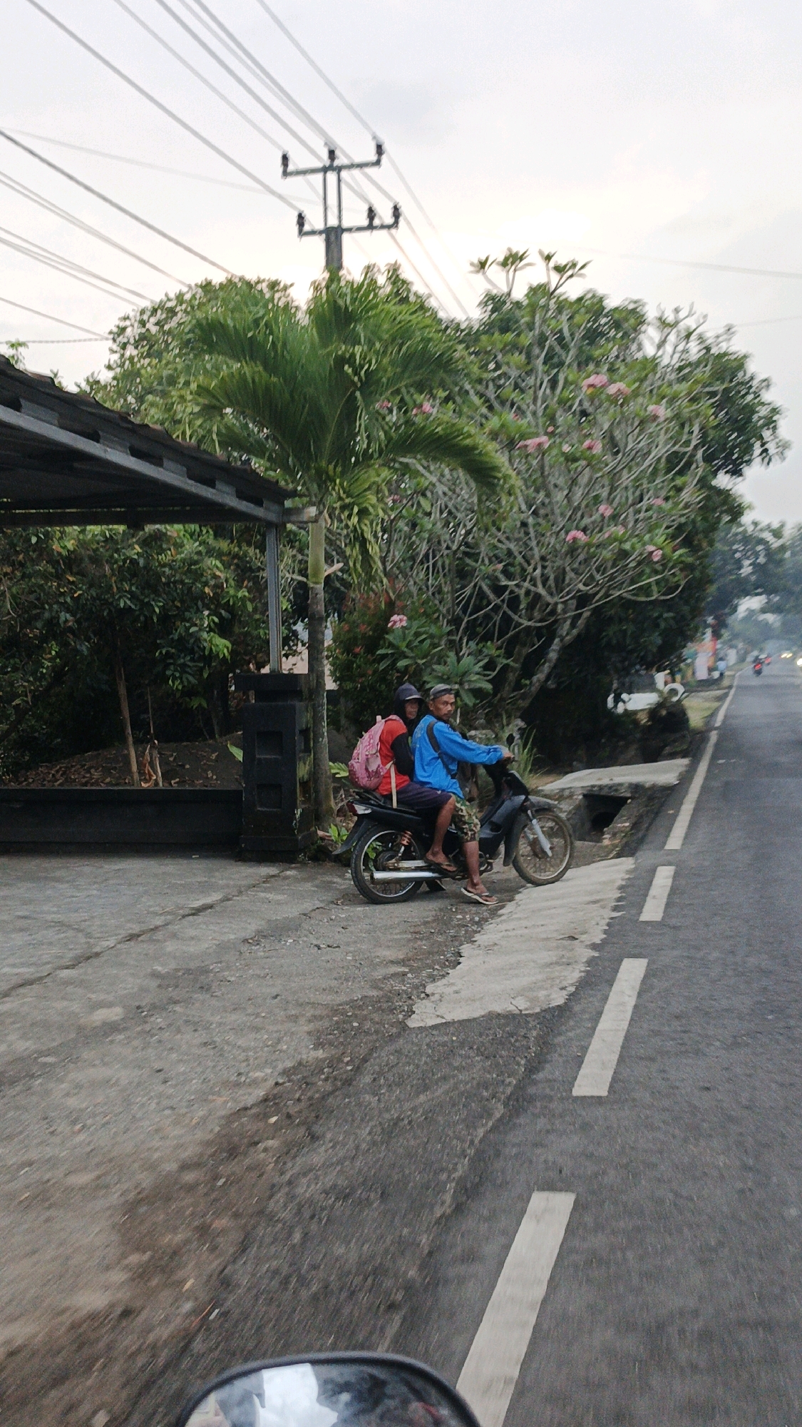 In Cibenda, Indonesia, two individuals are seen on a motorcycle by a road. The person in the rear, wearing a red backpack and a dark head covering, is facing away from the camera. The person in the front, wearing a blue long-sleeved shirt and patterned shorts, is looking forward and appears to be driving the motorcycle. They are positioned on the side of a paved road with a concrete gutter and a dirt area adjacent to it. In the background, there are trees, a large plant with fronds, and a utility pole with overhead wires. A dark structure with a sloped roof is visible on the left side of the image. The sky is overcast.