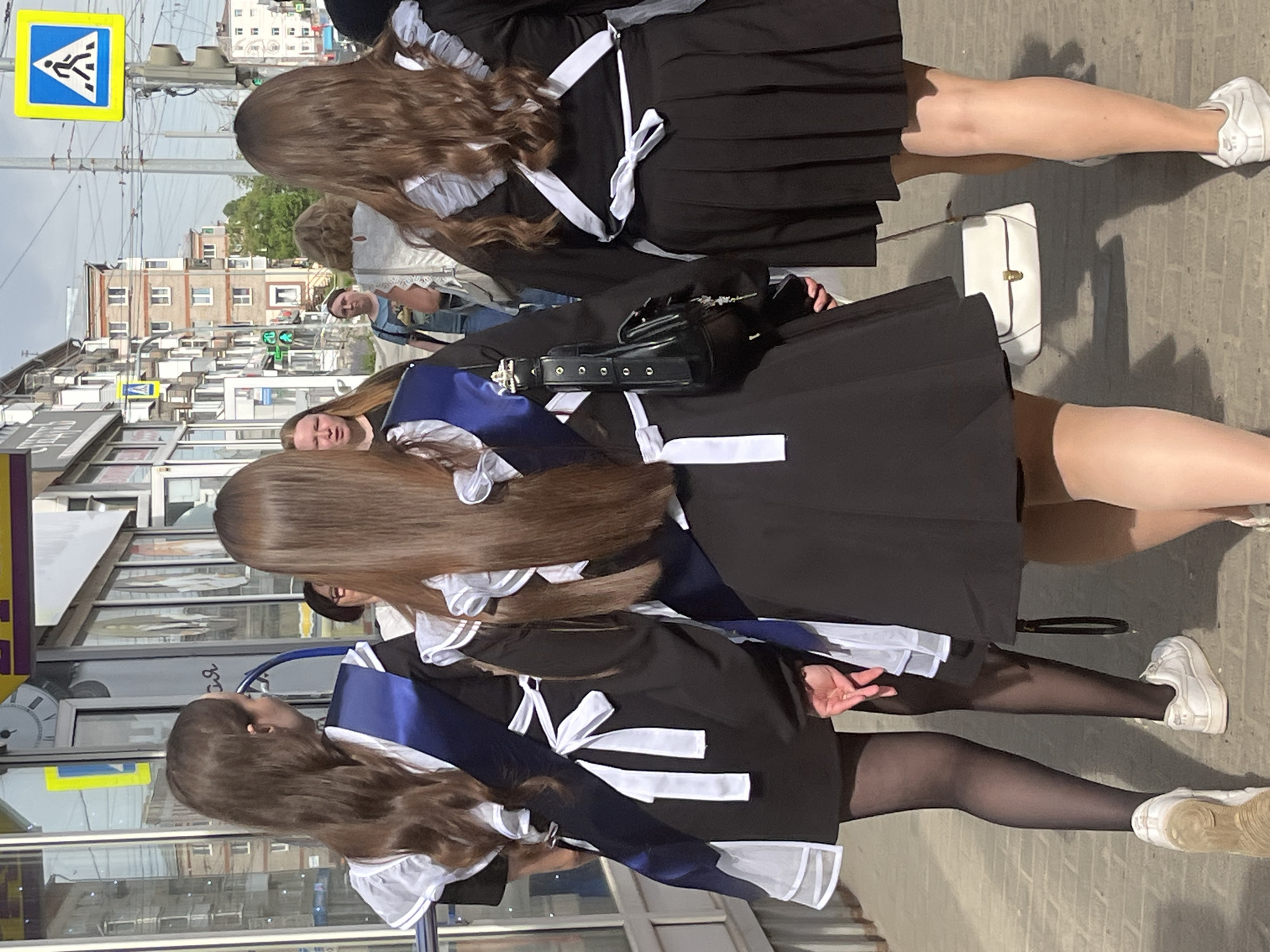 The image shows three young women standing on a sidewalk in Izhevsk, Russia. The women are dressed in matching black school uniforms with white accents and stockings. They are adorned with blue sashes, commonly worn by Russian high school graduates on their last day of school. The women are standing close together. The sidewalk is made of gray stone slabs, with a street visible in the background. A pedestrian crossing sign is visible in the upper left. Modern buildings are visible in the background. One woman has a small black purse with a silver buckle on her shoulder, and a white purse is near another one.